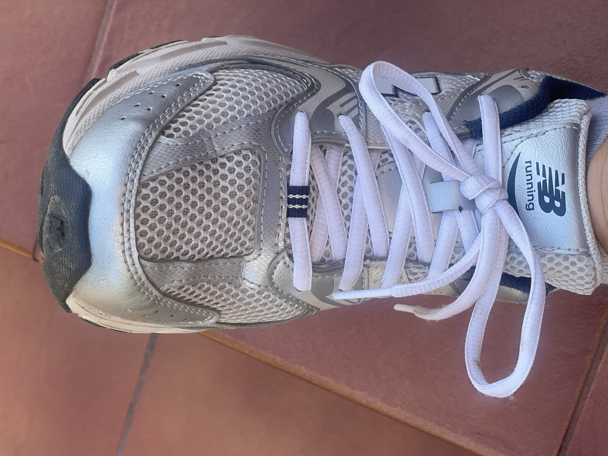 A close-up, top-down view shows the front of a silver and white New Balance athletic shoe resting on a reddish-brown tiled surface. The shoe features white laces tied in a knot, a mesh upper, and silver synthetic overlays. The New Balance logo and the word "running" are visible on a label on the tongue. The tiled surface appears to be outdoors, possibly a patio or walkway. There are no people visible in the frame.