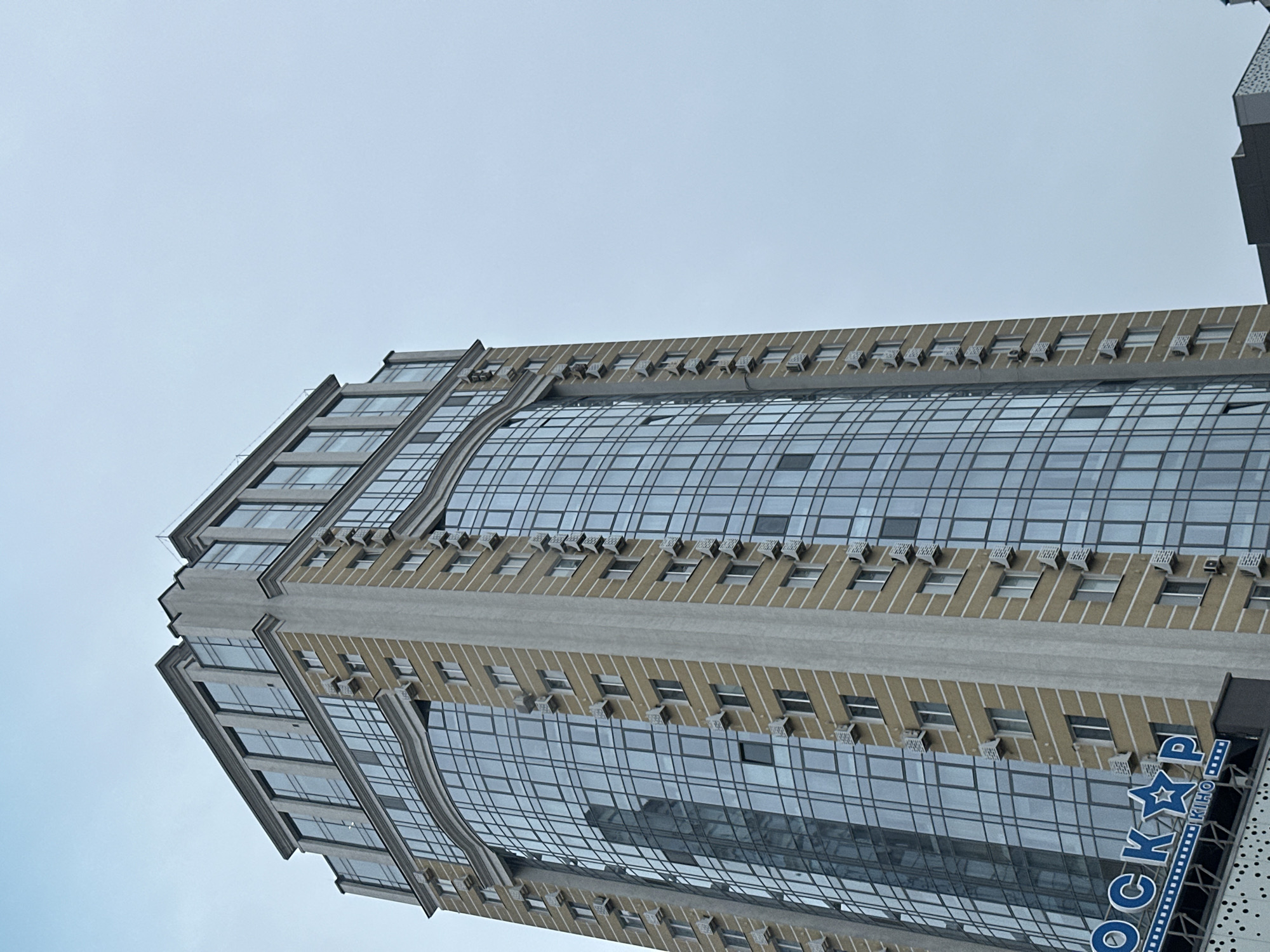 The image depicts a tall, modern building with a glass facade, likely located in Kyiv, Ukraine, based on the Cyrillic lettering on the building's sign which reads "Оскар кіно" (Oscar Kino). The building appears to be a cinema or entertainment complex, given the presence of the signage. The sky is overcast and the building is viewed from a low angle, emphasizing its height and imposing presence. The facade features regular rows of windows and architectural details that add texture to the structure.