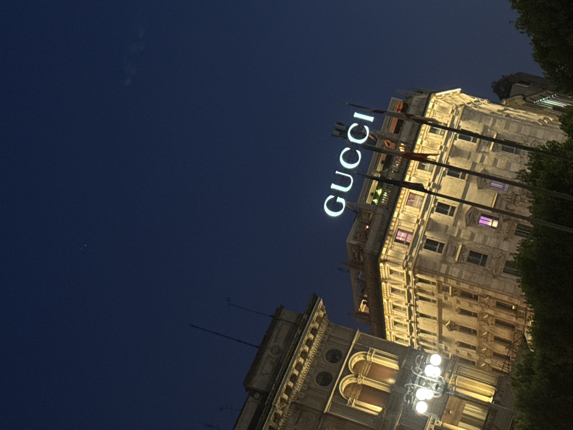 A low-angle shot presents the illuminated facade of a building at night. A brightly lit "GUCCI" sign is positioned atop the building. The architectural details of the building, including windows and ornate trim, are clearly visible. To the right, there is a patch of green foliage. The sky is dark blue. A smaller building with illuminated arches and a multi-bulb light fixture is at the bottom left. This is likely the Galleria Vittorio Emanuele II in Milan, Italy, recognizable by the Gucci sign on the building and the architecture of the Galleria. No people are visible.