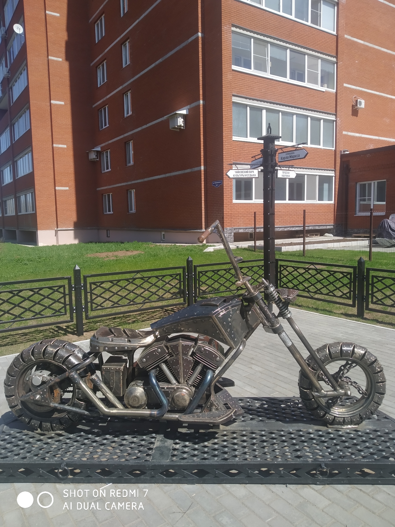 The image captures a detailed metal sculpture of a motorcycle situated in Chaikovsky, Russia, in front of a modern brick building. The sculpture features a rugged, industrial design with a visible engine and textured tires. A black metal fence encloses a grassy area in the background.
A signpost with directions to different locations in the city is visible; the text on the sign indicates directions to "Ploshchad Karla Marksa" (Karl Marx Square), "Chaykovskiy Park Kultury I Otdykha" (Chaikovsky Park of Culture and Recreation), and "Gorodskaya Naberezhnaya" (City Embankment).