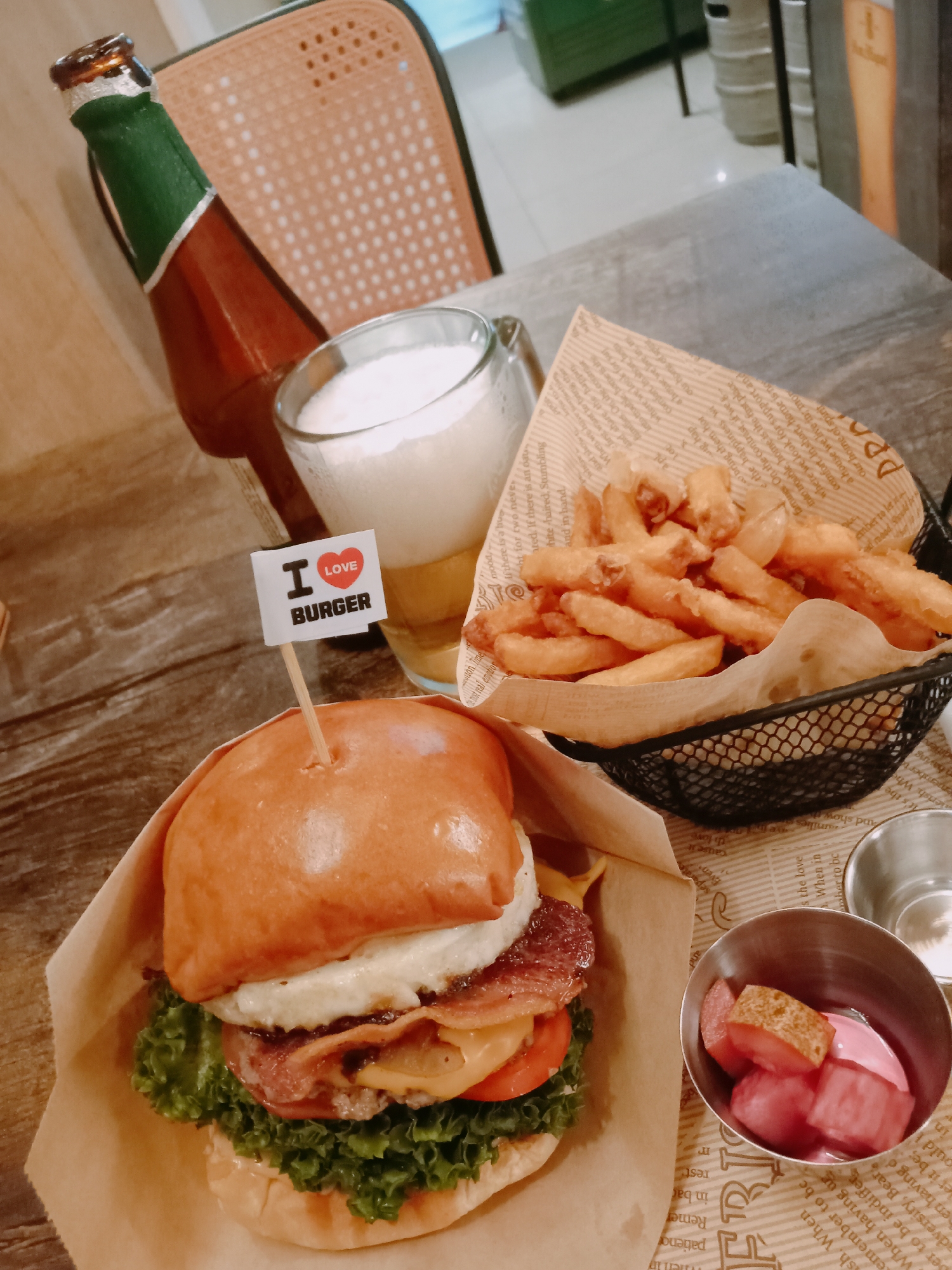 A tabletop scene in Nha Trang Ward, Vietnam, features a burger, fries, beer, and pickled vegetables. A craft beer bottle stands next to a glass of foamy beer. A burger with lettuce, tomato, cheese, bacon, and a fried egg is displayed with an "I Love Burger" toothpick. French fries fill a black wire basket. Pickled vegetables sit in a small metal bowl. All items are placed on paper with printed text. A wooden chair is visible in the background, and a white barrel-like object is in the background.