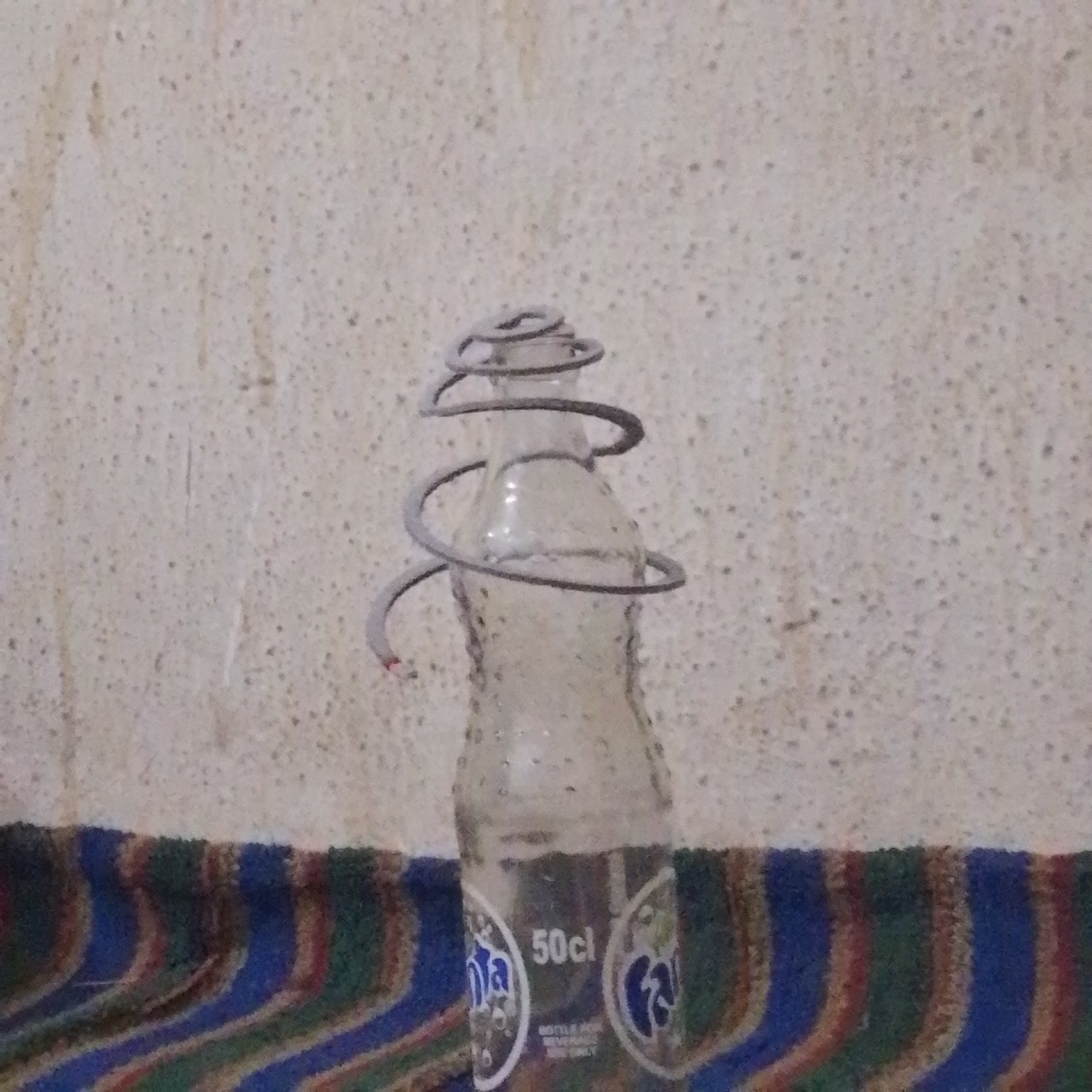 A clear glass bottle of Fanta soda sits on a striped cloth in front of a textured wall. A coiled metal object is wrapped around the neck of the bottle, with the bottom end appearing to be lit or smoldering. The bottle has labels indicating "50cl" and the Fanta logo. The scene appears to be indoors, likely in a residential setting in Maiburiji, Nigeria.