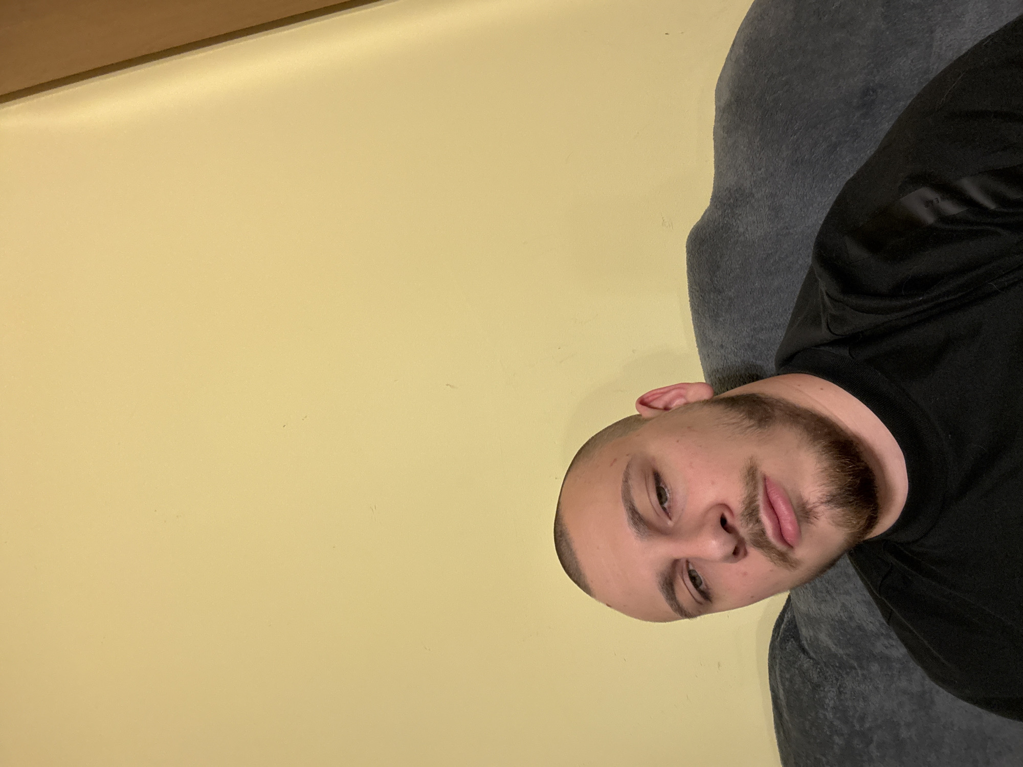 A man with a shaved head and a beard is looking at the camera. He is wearing a black shirt. He is sitting in a gray chair. The background is a yellow wall. The location is unknown.