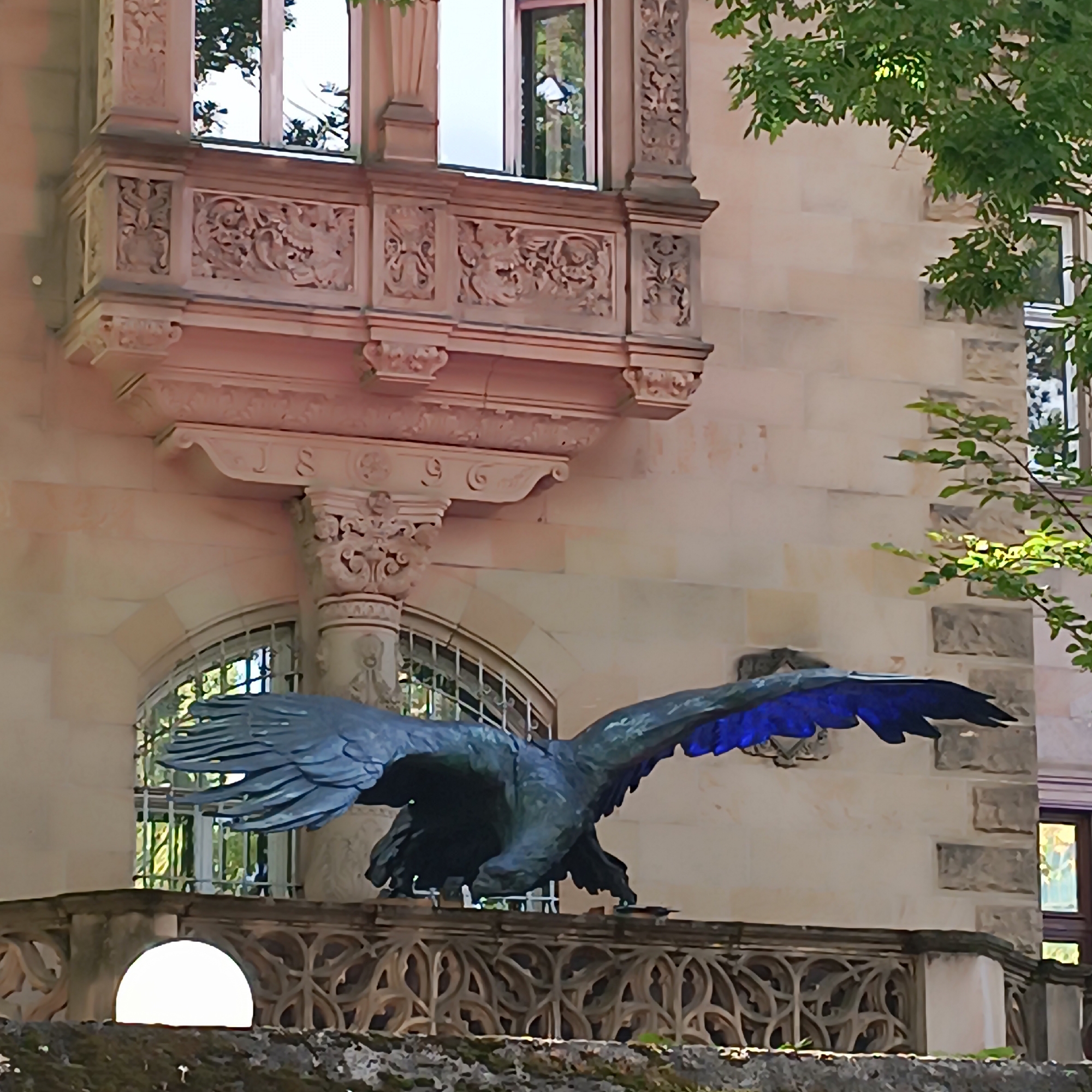The image shows a close-up view of a weathered, dark-colored eagle sculpture with blue accents on its wings, perched on a stone balcony railing in front of a building with ornate architectural details. The building's facade is constructed from light-colored stone blocks, and it features decorative carvings and multiple windows, some of which reflect greenery from surrounding trees. The balcony itself is elaborate, with intricate stonework and a patterned railing. The overall impression is of an old, possibly historical building, with a prominent sculptural element on its exterior. The specific location is not identifiable from the image alone, but it is presented in the context of Frankfurt, Germany. There are no people visible in the image.