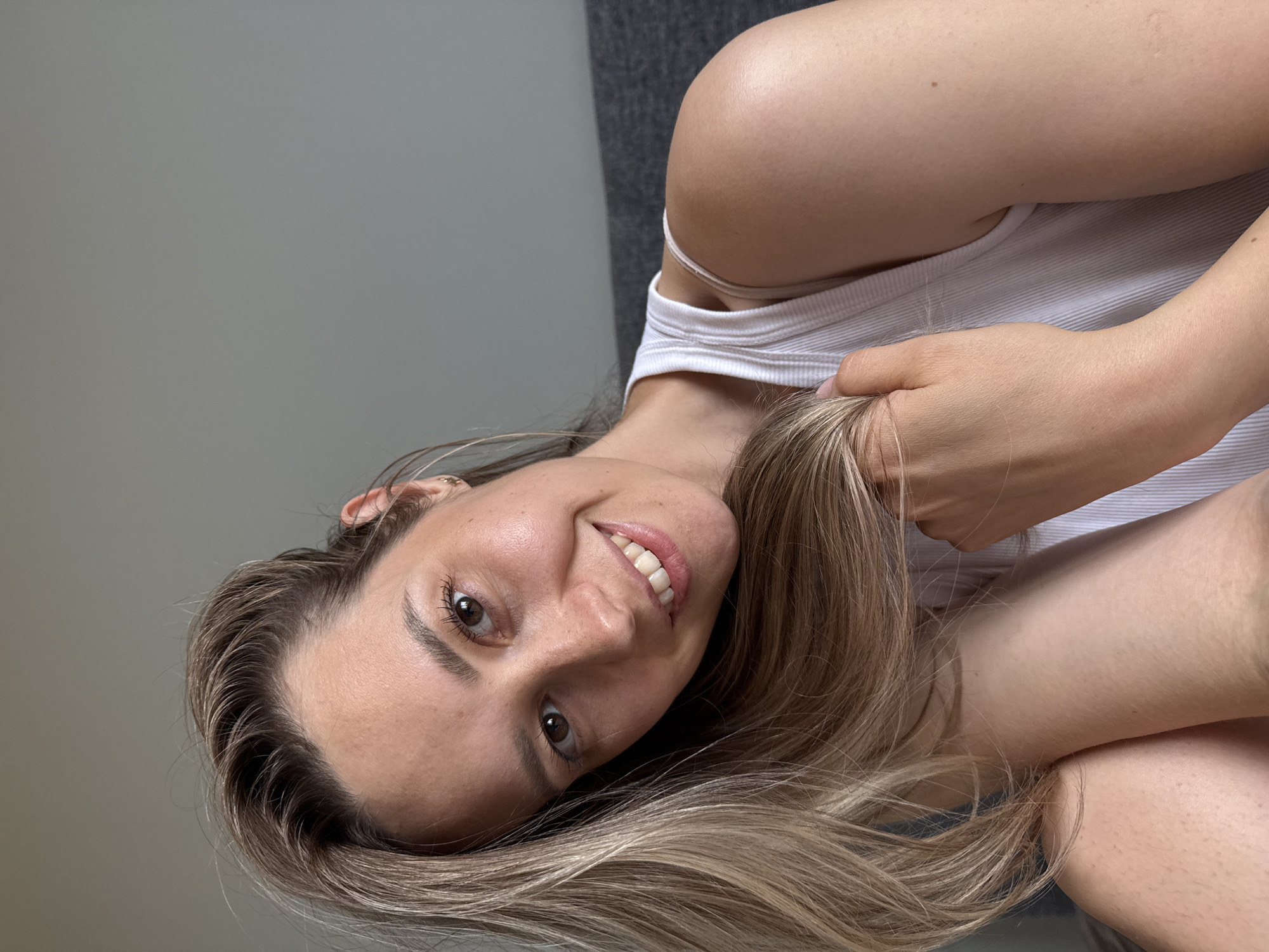 A young woman is pictured indoors. She is fair-skinned with light brown eyes and blonde hair. She is wearing a white ribbed tank top. She is smiling, and part of her hair is in her hand. The background consists of a grey wall and a section of grey fabric (possibly a couch). There's no visual information to suggest the location is Chaikovsky, Russia.