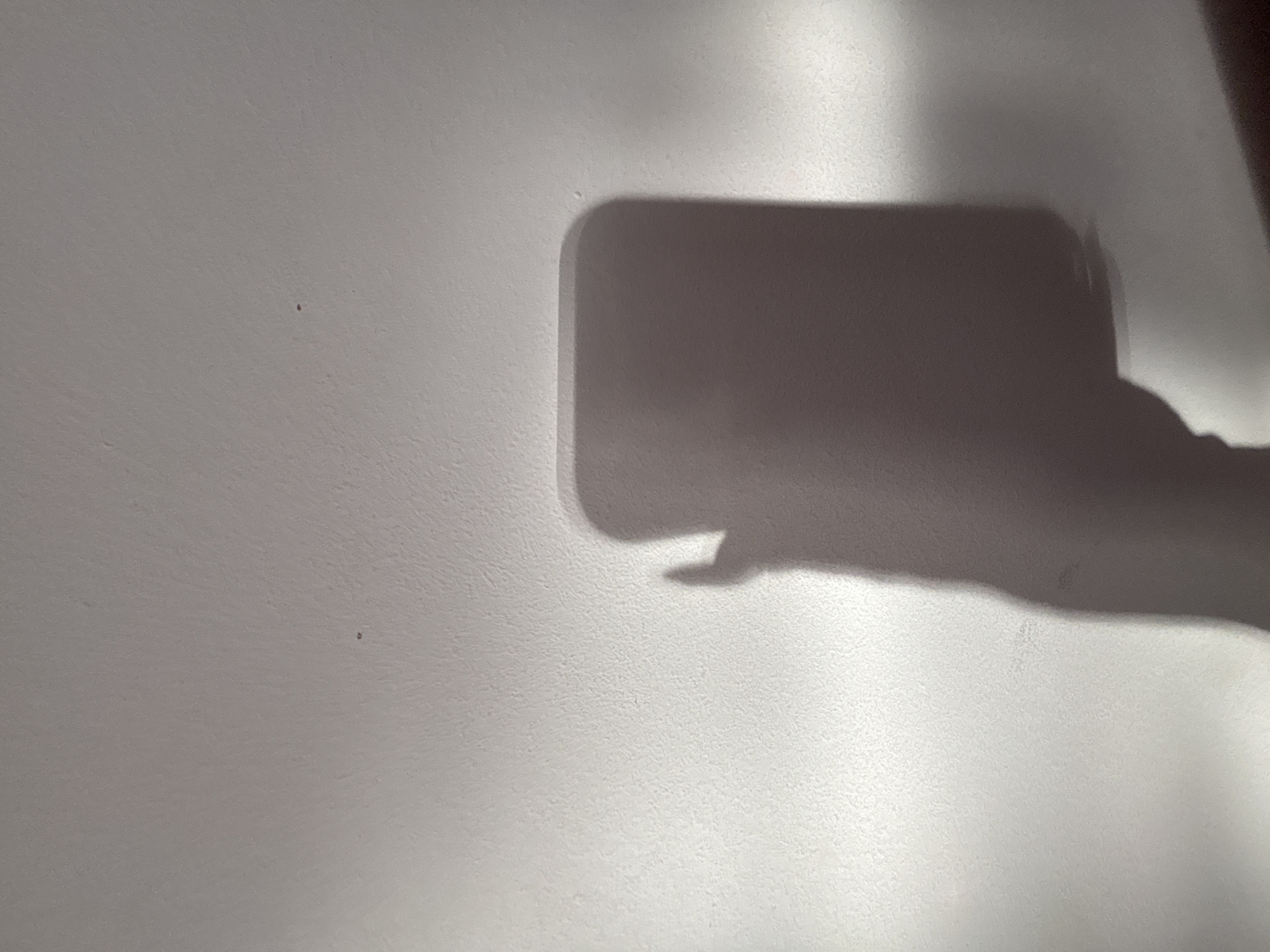 The image shows a white wall with shadows cast on it. The shadows appear to be of a hand holding a rectangular object, possibly a phone. The location in the city of Troyan, Bulgaria is not discernable from the image.