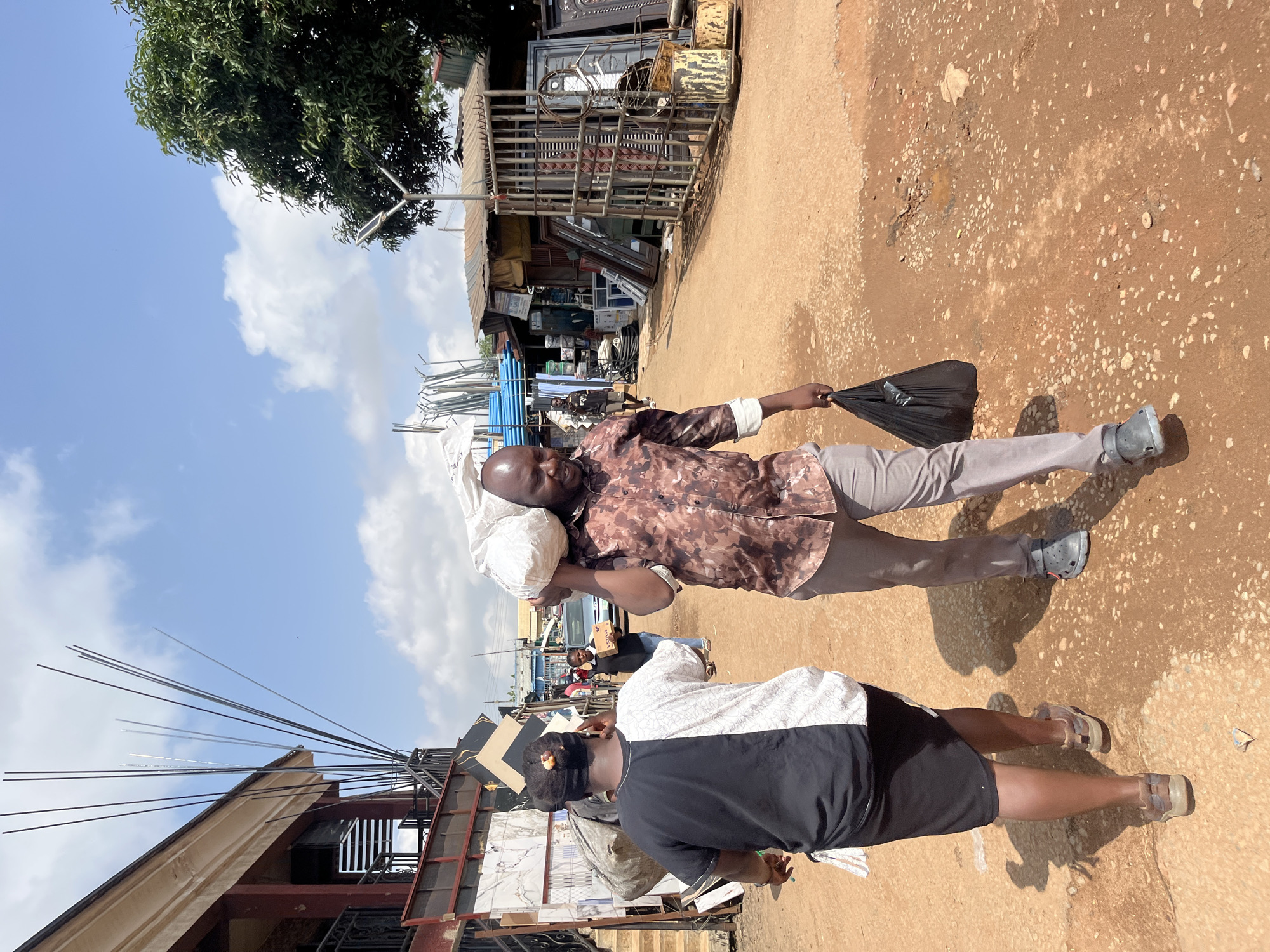 A man, possibly in his 30s or 40s, is carrying a large white bundle on his shoulders and a black plastic bag in his right hand. He is walking on a dirt path. He is wearing a camouflage patterned shirt and light-colored trousers. The woman in front of him is wearing a black and white top and dark shorts. She is also walking on the dirt path, holding something in her hands. In the background, there are shops with metal structures and various building materials. The scene appears to be an outdoor market. The sky is blue with some white clouds, suggesting it is daytime and the weather is fair. The overall setting appears to be a busy marketplace in Gura Top, Nigeria, with commercial activities taking place. The man's expression is not clearly visible, but his posture suggests he is engaged in carrying his load. The woman's face is obscured.