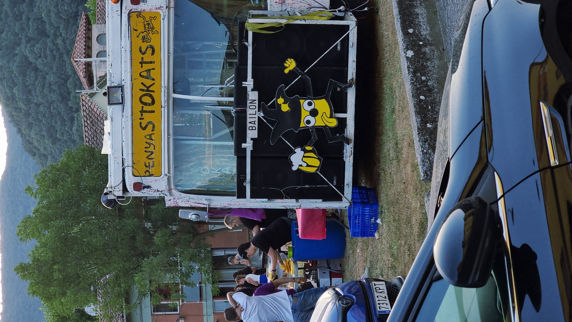 This image captures a street scene in Sant Joan Les Fonts, Spain, likely during an outdoor event or festival. In the foreground, a dark blue car's hood and side mirror are visible, with a portion of a license plate reading "7312 KPT." Behind the car, a white food truck or mobile vendor is the central focus. The truck is adorned with a yellow sign that reads "PENYAS'TOKATS" in black lettering, and features a whimsical illustration of a yellow character wearing a jester's hat and holding a mug of beer, with its tongue sticking out. The truck is also equipped with large black speaker boxes on its rear.
Several people are present around the truck, appearing to be interacting or serving food. One person in a white t-shirt and jeans is standing on the left, facing away from the camera, with their hand on their head. Others are gathered behind a table laden with food items, suggesting a food service operation. The environment appears to be a grassy outdoor area with trees and buildings in the background, and a hilly, tree-covered landscape visible beyond the buildings, characteristic of the region. The lighting suggests it is late afternoon or early evening.