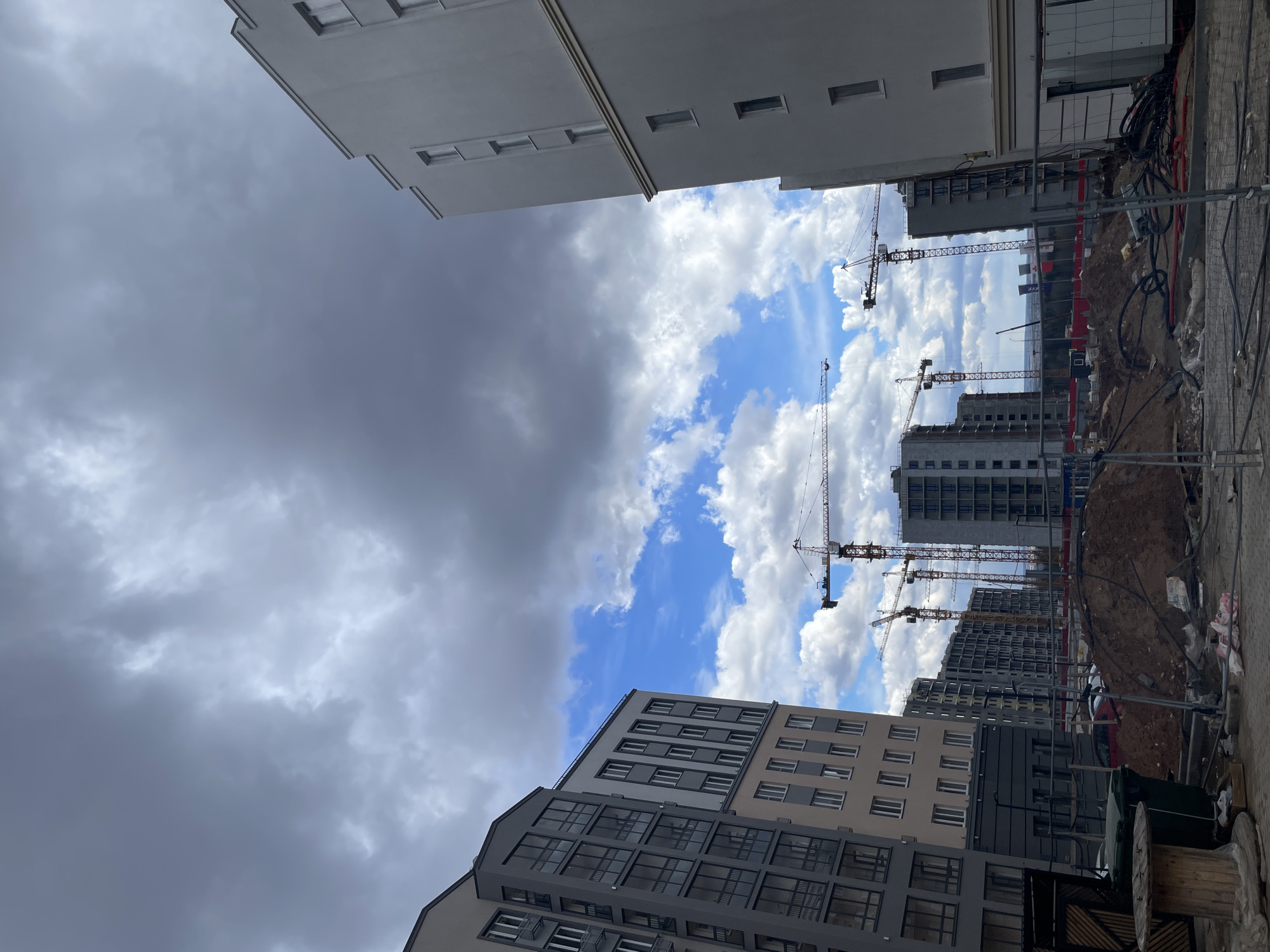 The image captures a low-angle perspective looking up at an urban construction site, framed by multi-story buildings, likely in Pervomaysky, Russia. The sky presents a striking contrast: the upper left portion is dominated by heavy, dark grey, overcast clouds, while the lower right reveals a vibrant blue sky dotted with bright, fluffy white cumulus clouds.
In the foreground and background, various modern residential or commercial buildings are visible. Some appear largely complete with clean facades in shades of white, light brown, and dark grey, featuring numerous rectangular windows. Others are in earlier stages of construction, showing exposed concrete and scaffolding.
The central and right-hand side of the image prominently features an active construction site. Multiple tall tower cranes extend upwards, indicating ongoing building work. Piles of reddish-brown earth, rebar, construction fences, and bundled cables or hoses are scattered across the site. A portion of a red car is discernible amidst the construction materials. In the immediate foreground, a large wooden cable spool is visible.
On a white banner or sign within the construction zone, the Cyrillic word "МАРК" (MARK) can be partially seen. The scene depicts daytime conditions with distinctly mixed weather, ranging from bright sunshine to impending overcast skies, characteristic of a developing area undergoing significant construction.