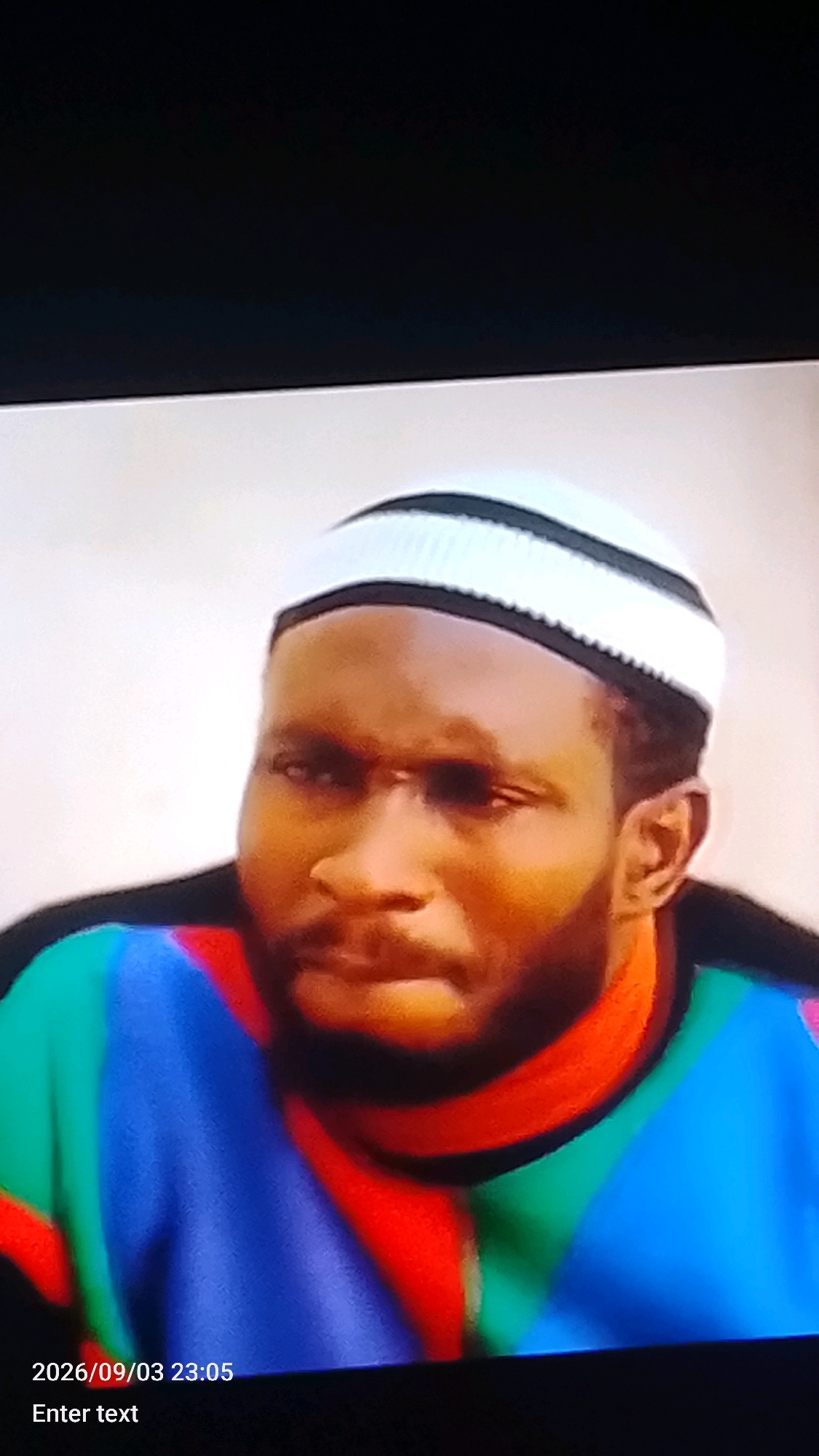 The image shows a close-up of a man wearing a white and black striped kufi cap and a colorful shirt. The shirt has blocks of red, blue, and green. The man has a beard and appears to be looking slightly off-camera. In the bottom left corner, a timestamp "2026/09/03 23:05" is visible, along with the text "Enter text". The background is a plain, light color, possibly a wall. There is no specific context or setting within the city of Lokoja, Nigeria, that can be identified from this image.