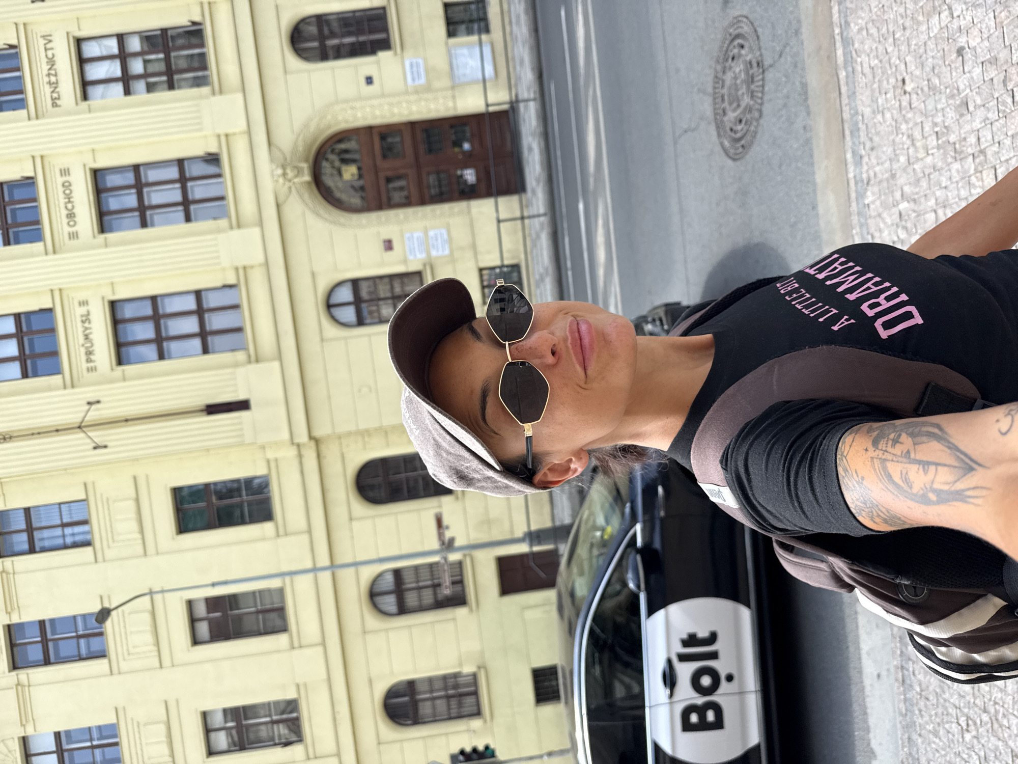 A woman with dark hair, wearing a baseball cap, sunglasses, and a black t-shirt with pink text that reads "A LITTLE BIT DRAMA," walks down a street in Prague, Czechia. She has a backpack on and a tattoo on her left arm. A black Bolt car is parked behind her on the street. The building behind her is light yellow with ornate architectural details and several windows. The street is paved with cobblestones.