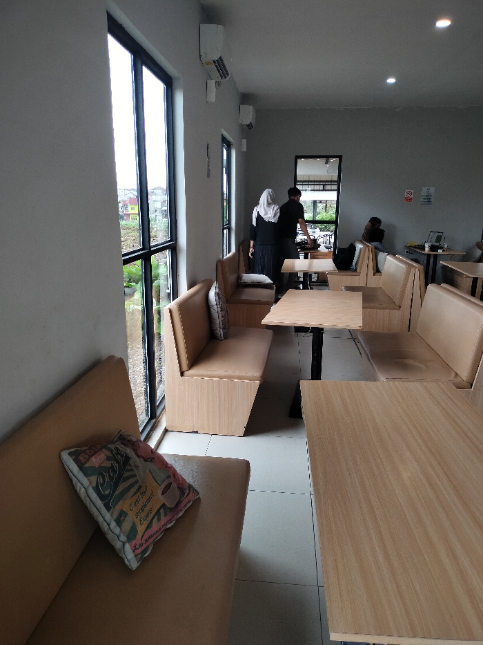 The image depicts the interior of a modern cafe or restaurant in Dramaga, Indonesia, characterized by a minimalist aesthetic. The walls are light gray, and the floor is tiled in white. Large, floor-toceiling windows line the left side, offering a view of a green, outdoor landscape with trees and distant residential buildings, including a distinct yellow house.
The cafe is furnished with booth-style seating. Light wood benches with beige upholstered cushions are arranged around light wooden tables with dark metal bases. In the foreground, one cushion features a decorative print with French text, including "Cafe" and "C'est tout simplement Expres," along with an illustration of a coffee cup.
Several individuals are present in the cafe. In the mid-ground, two people are standing near a counter or table: one wearing a white hijab and dark clothing, and another in a dark t-shirt. They appear to be interacting or attending to something. Further back on the right, a person is seated at a booth with a laptop on the table.
The ceiling features recessed lighting, and multiple white wall-mounted air conditioning units are visible on the gray walls. A "No Smoking" sign is also visible on the far wall. The natural light flooding through the large windows suggests it is daytime. The overall atmosphere appears calm, with a few patrons and staff in the space.