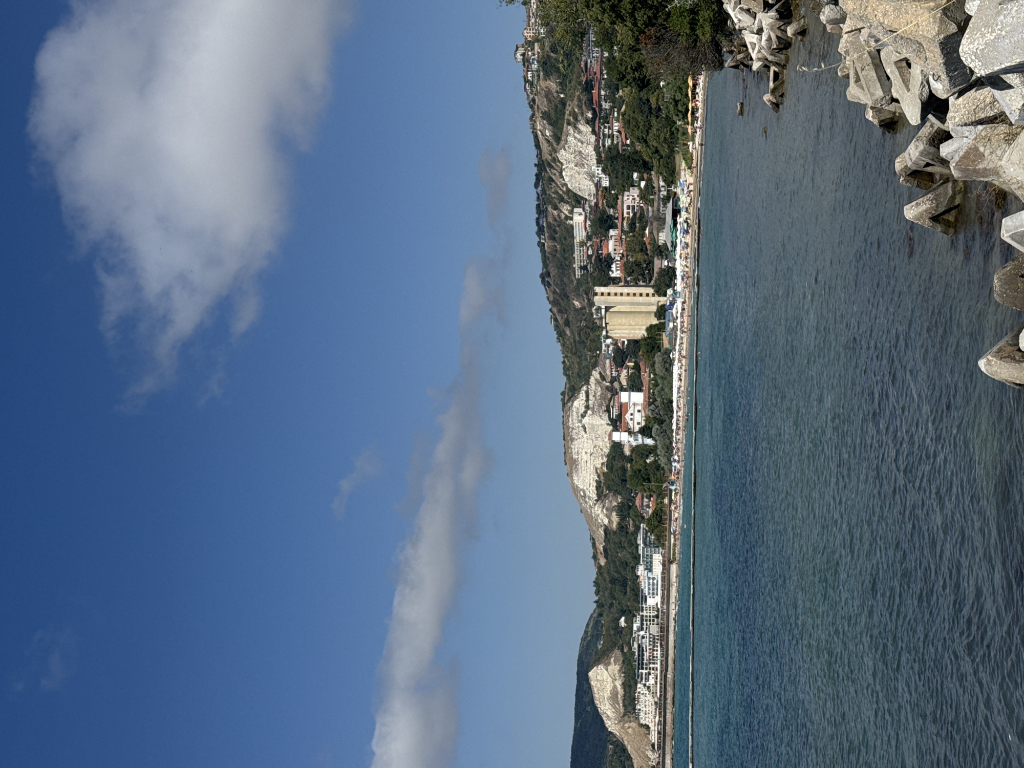 This is a wide shot of the coastline in Balchik, Bulgaria, on a bright, sunny day with a clear blue sky. In the foreground, the dark blue water is calm with slight ripples. Along the right edge of the frame, a jumble of large, gray, concrete breakwater blocks are visible. A thin, horizontal line stretches across the water, likely a barrier or another breakwater.
In the middle ground, a wide sandy beach is dotted with numerous umbrellas and beachgoers. Behind the beach, a cluster of white buildings, possibly hotels or apartment complexes, runs along the shoreline. Further back, a town is built into the hillside, with a variety of buildings, including some with red roofs. The hills themselves are a mix of green vegetation and lighter-colored, rocky slopes. A tall, cylindrical building, a light-colored silo, stands out among the town's structures.
The sky is a vibrant blue with scattered white clouds. The overall impression is a serene coastal scene with a bustling beach area and a town nestled into the surrounding landscape.