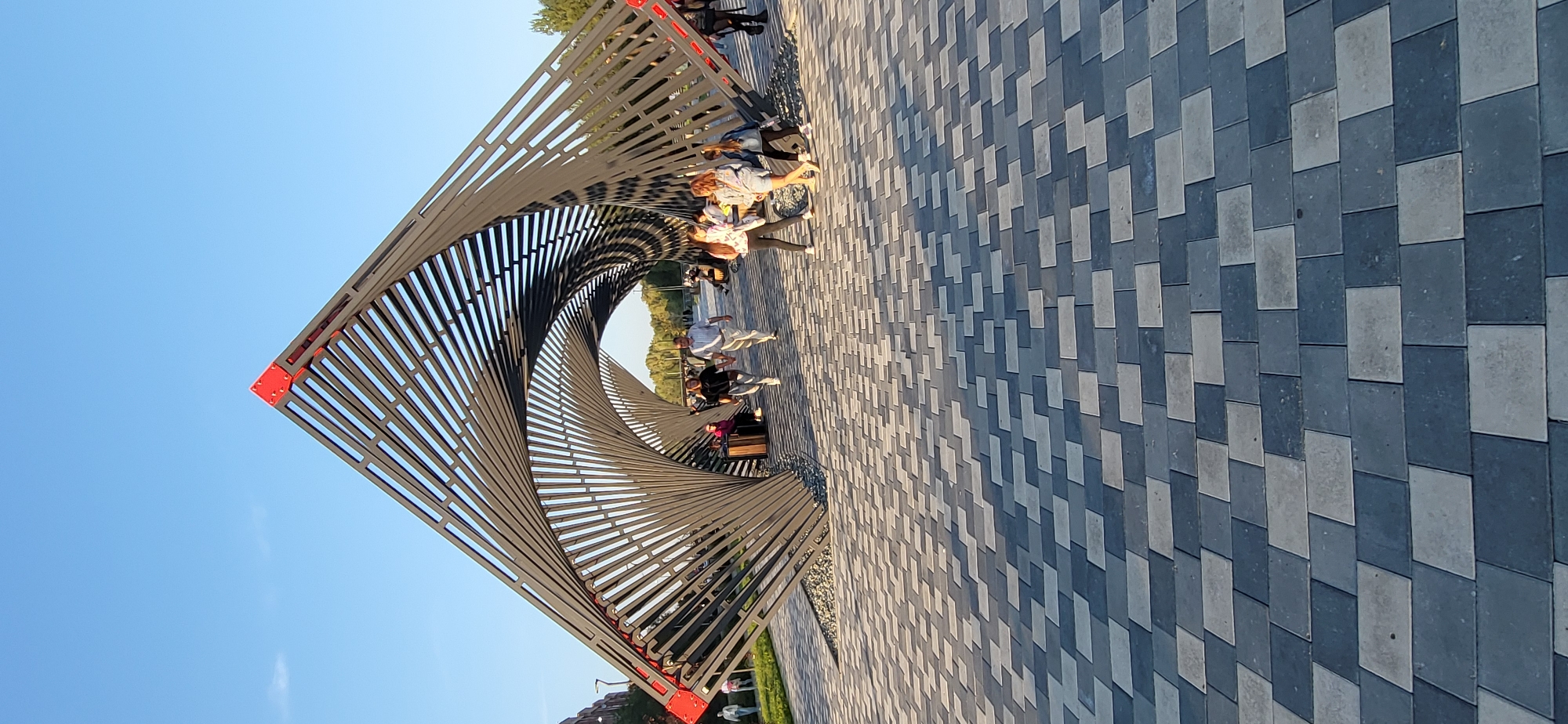In Oskemen, Kazakhstan, a modern park features a striking sculptural installation resembling an open tunnel or gateway. The structure, made of dark metal beams arranged in a spiral, is adorned with vibrant red accents at its apex. The ground is paved with a mosaic of rectangular stones in shades of gray and dark blue, creating a geometric pattern. Several individuals are present, with some walking through the structure and others seated nearby. A couple walks hand-in-hand through the archway, while a group of young people are gathered to the right, some seated. In the background, lush green trees and a clear blue sky suggest a pleasant day. The scene captures a blend of contemporary art and public space, inviting visitors to explore and relax.
