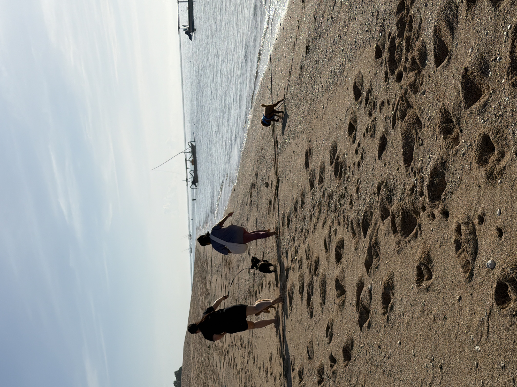 The image depicts a beach scene in Unknown, Indonesia. The camera is oriented horizontally, showing the shoreline running from the upper left to the lower right. The sky is light and bright, with a hint of clouds. The sea is calm with several boats in the distance. In the foreground, the sand is textured with footprints. Three people are visible: one person is crouching near the water's edge, and two people are walking away from the viewer, with a dog on a leash.