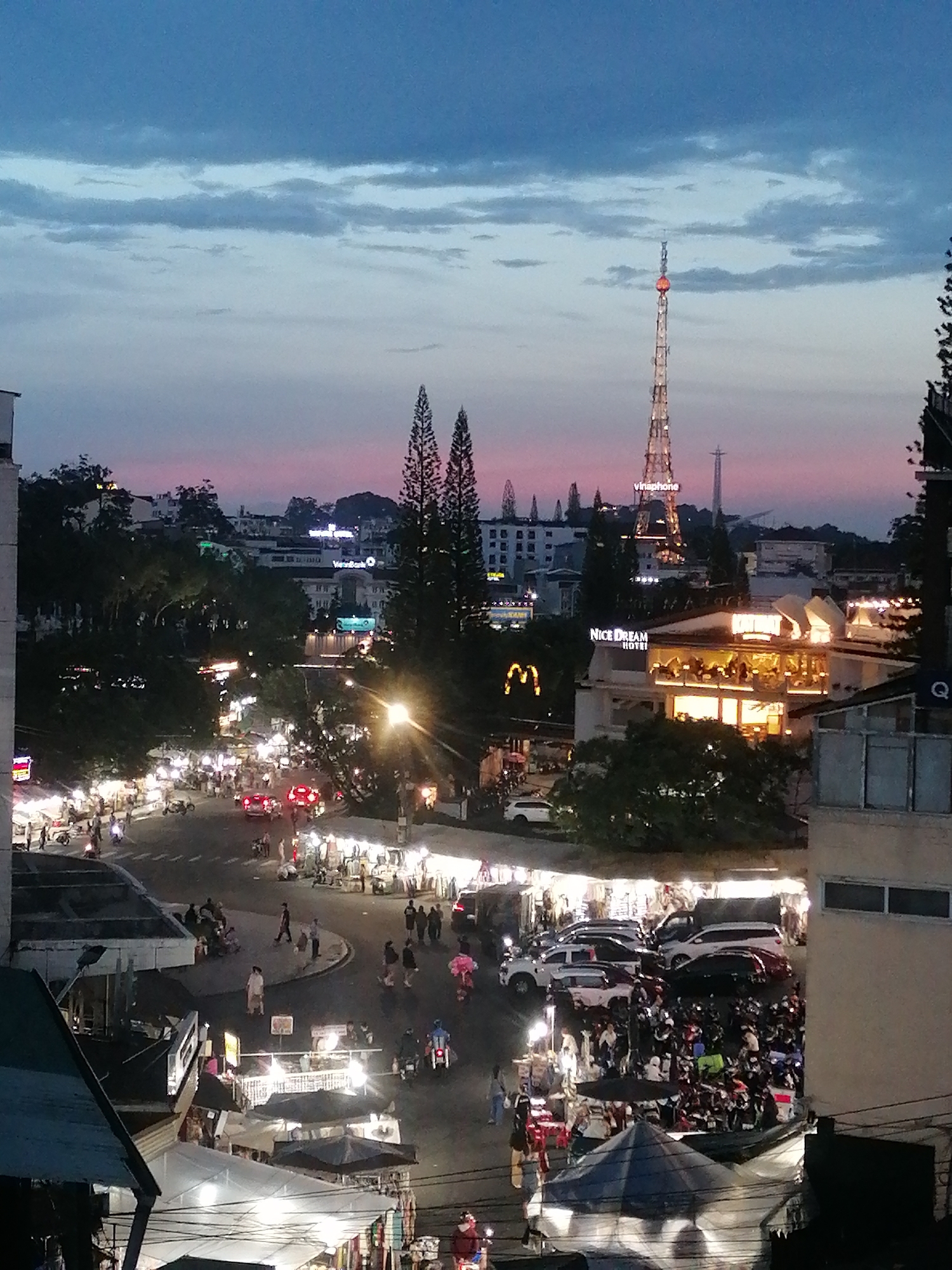 The image depicts an elevated, nighttime view of Da Lat, Vietnam. A bustling city street is visible, filled with pedestrians, motorcycles, and a few cars, all illuminated by streetlights and storefronts. Several vendor tents line the street. In the mid-ground, a hotel named "Nice Dream Hotel" can be seen, along with a McDonald's restaurant. A tall telecommunications tower, labeled "vinaphone," rises above the cityscape. The sky is a blend of blue and pink hues, suggesting twilight. Buildings are visible in the background, indicating a moderately dense urban environment.