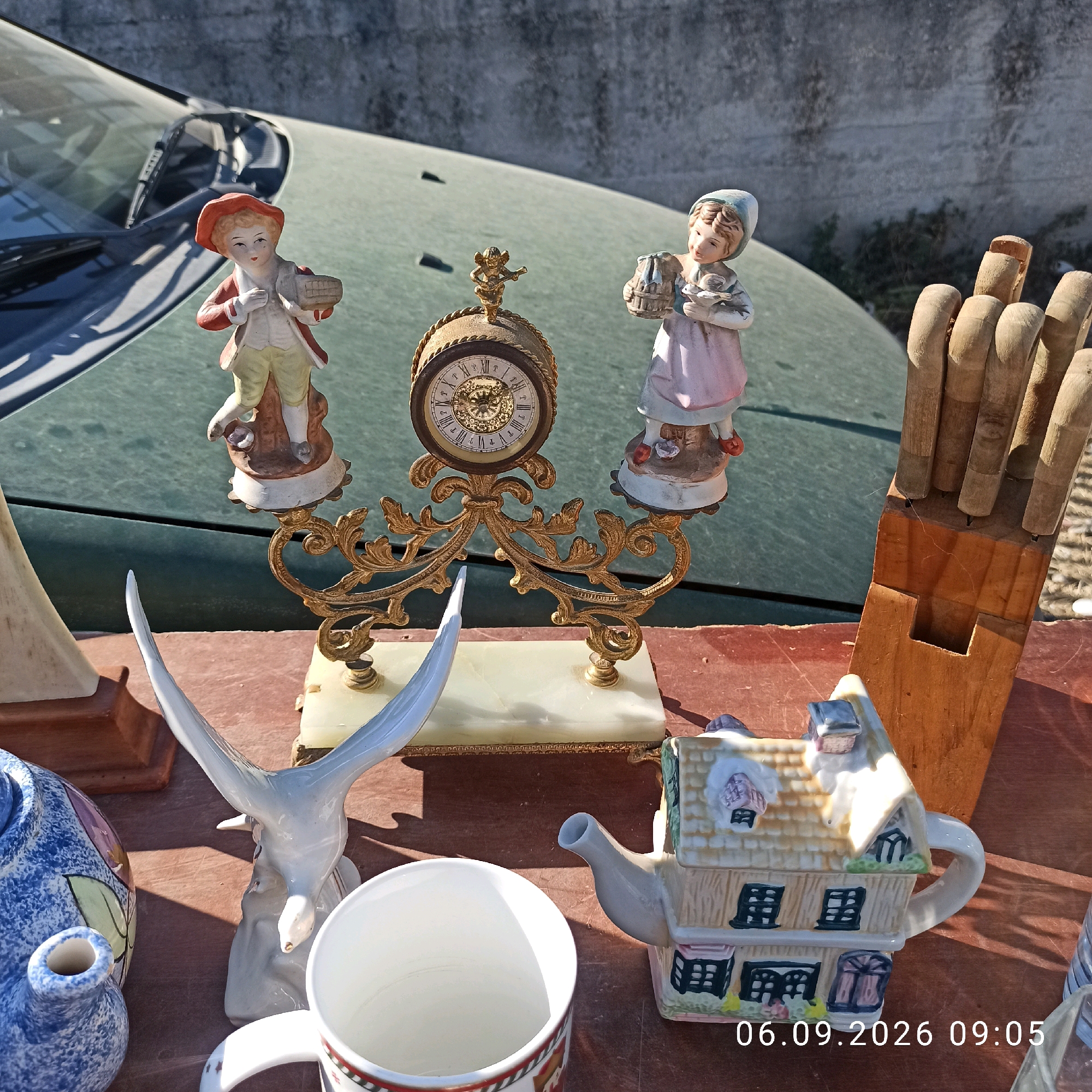 An outdoor market stall in Sannicandro di Bari, Italy, displays various decorative items. In the background, a dusty green car is parked. In the foreground, on a reddish-brown surface, are several ceramic figurines: a boy in a red hat and waistcoat, a girl in a pink dress, and a decorative clock with a cherub on top. To the right, a wooden block holds several wooden handles. In the lower part of the image, a blue and white patterned teapot, a white ceramic bird figurine, and a white mug are visible. To the right of the mug is a whimsical teapot shaped like a multi-story house. A timestamp "06.09.2026 09:05" is overlaid on the bottom right of the image.