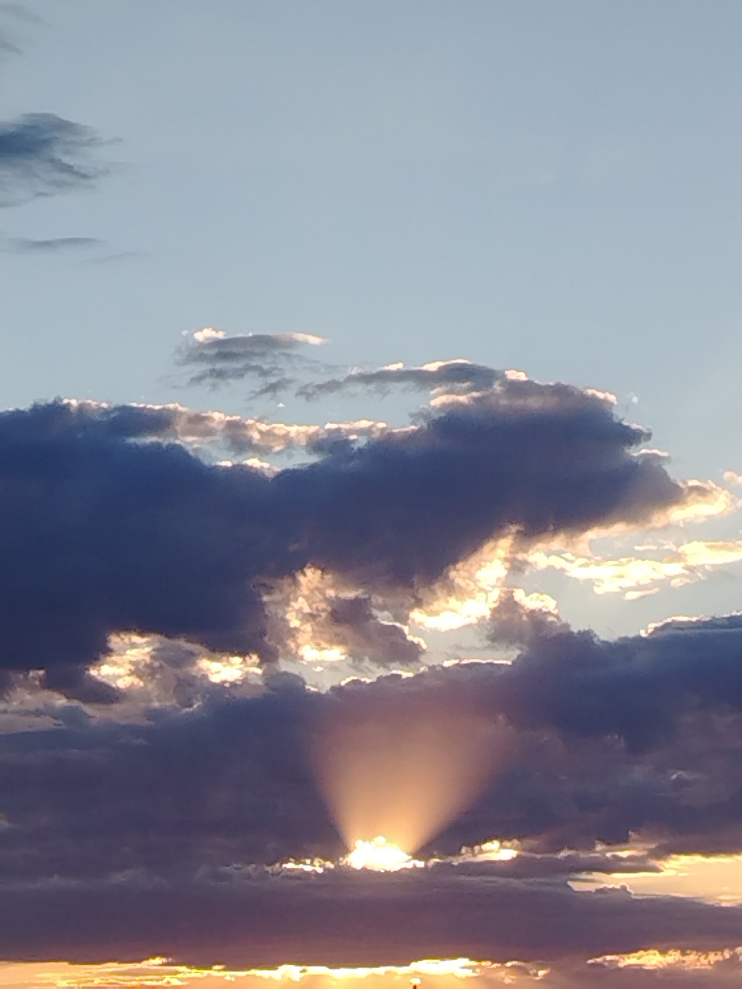 A vertical, medium shot captures a dramatic sunset over Baikonur, Kazakhstan. The sky is a pale blue, fading to a warmer yellow-orange at the horizon, where the sun's rays are just visible through a bank of dark, textured clouds. These clouds are illuminated from behind, creating a halo effect and casting a warm glow. In the center of the frame, a distinct sunbeam radiates outwards from behind a thick cloud mass, creating a crepuscular ray effect. The clouds themselves vary in color from deep indigo to lighter shades of grey and purple, with the edges highlighted by the setting sun. The lower portion of the image shows a sliver of land, possibly a building or structures, silhouetted against the vibrant horizon. No people or animals are visible.
