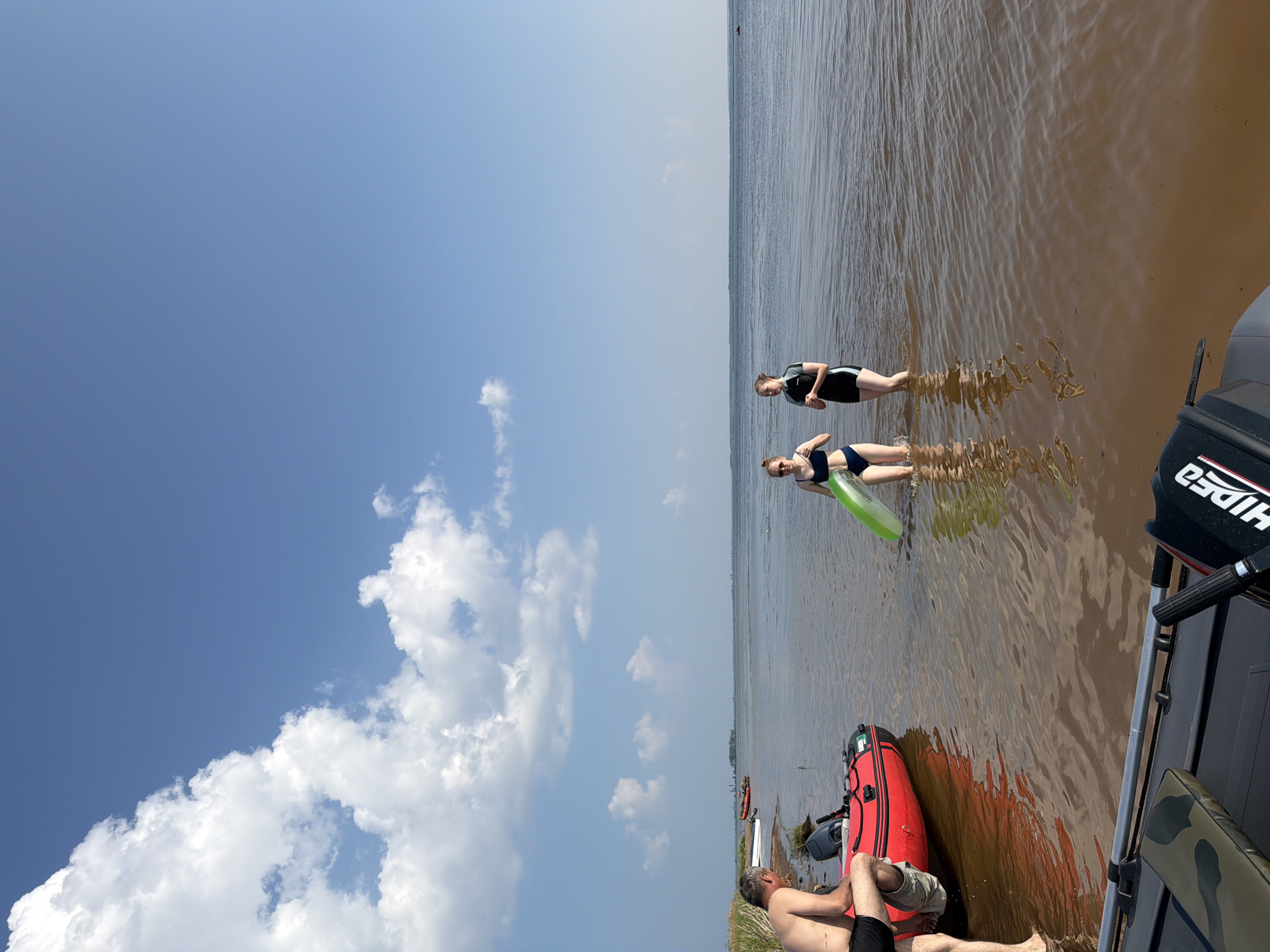 In Krasnyi Bor, Russia, on a sunny day with scattered clouds, a man is seen sitting on the edge of a red inflatable boat, partially submerged in murky water. Two young women are wading in the water near a large body of water. The woman on the left is wearing a blue bikini and holding a green inflatable ring. The woman on the right is wearing a black and white wetsuit. Another inflatable boat, partially in the foreground, is visible. In the distance, the horizon shows a calm sea or large river, with a few distant boats and landmass. The overall scene depicts a recreational activity by the water.