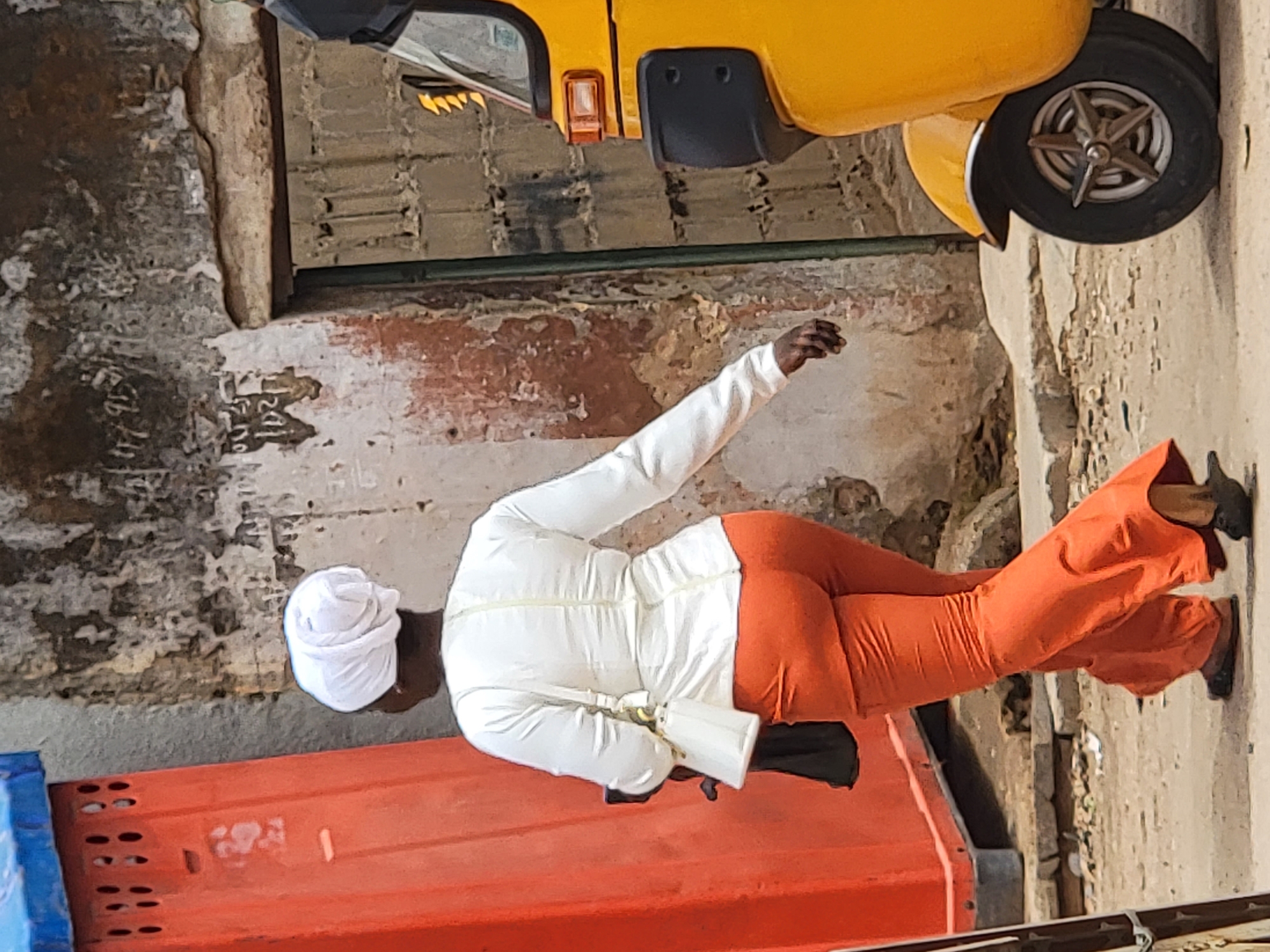 This image captures a bright daylight scene in Maiburiji, Nigeria, depicting a person walking away from the camera on what appears to be a street or alleyway. The lighting is harsh and bright, indicative of midday sun.
The central subject is a person, viewed from behind, dressed in a long-sleeved white top, loose-fitting bright orange trousers, and a white turban-style head covering. They are wearing dark shoes, and a white bag or pouch is visible across their body. Their right arm is slightly raised and bent, suggesting motion.
In the upper part of the frame, a bright yellow three-wheeled vehicle, resembling an auto-rickshaw or tuk-tuk, is partially visible. Its front wheel, a portion of its body, and a rearview mirror are distinguishable.
The setting is characterized by weathered infrastructure. The ground is an uneven, dusty surface. The walls on either side of the scene are old and show significant signs of wear, with peeling paint or plaster, exposed brickwork, and various textures. Faint, illegible markings or graffiti can be seen on the wall to the left. In the lower left foreground, a section of a large, bright orange container or barrier with a blue top is visible.
The scene conveys an everyday urban or semi-urban moment, with the person seemingly engaged in their daily commute or activity. No specific emotions are discernible from the person's back.
