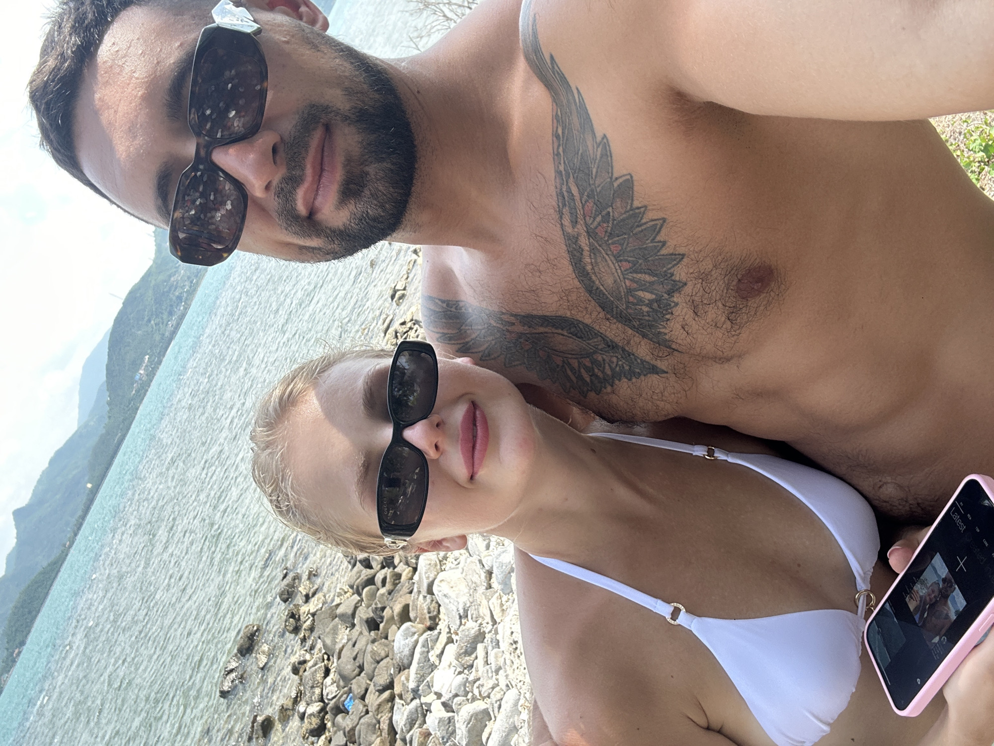 A man and a woman are taking a selfie on a rocky beach. The man is shirtless, revealing a large tattoo on his chest. The woman is wearing a white bikini top. The ocean is visible behind them, with mountains in the distance. The man is wearing dark sunglasses, and the woman is wearing black sunglasses. The woman is holding a pink smartphone. The location is the city of Ninh Ích, Vietnam.