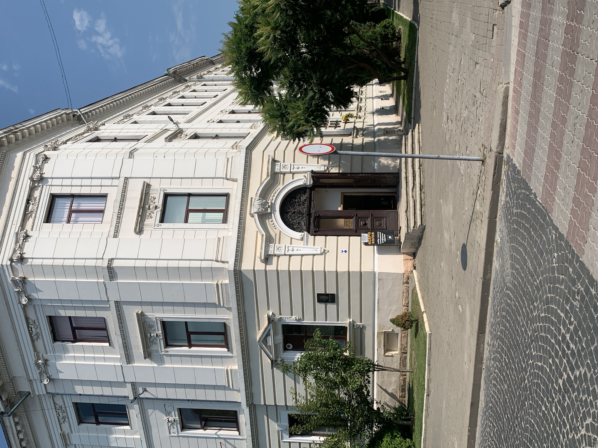 The image shows the corner of a white, multi-story building with ornate architectural details. The building has numerous windows with dark frames. In front of the building, there is a paved sidewalk and a street made of cobblestones. A pedestrian crossing with red and white stripes is visible. There are trees and some greenery around the building. A sign with a red circle and a horizontal white bar is mounted on a pole in front of the building. The scene appears to be a street in a city, likely during the daytime, under a clear blue sky. The building is located at the intersection of vul. Soborna and vul. Vovchynetska in Kolomyia, Ukraine. The ground floor of the building has an entrance with a dark wooden door and steps leading up to it. Above the entrance, there is an arched decorative element. A sign with Cyrillic text, "КСЕРО КОПІЯ," is visible to the right of the entrance.