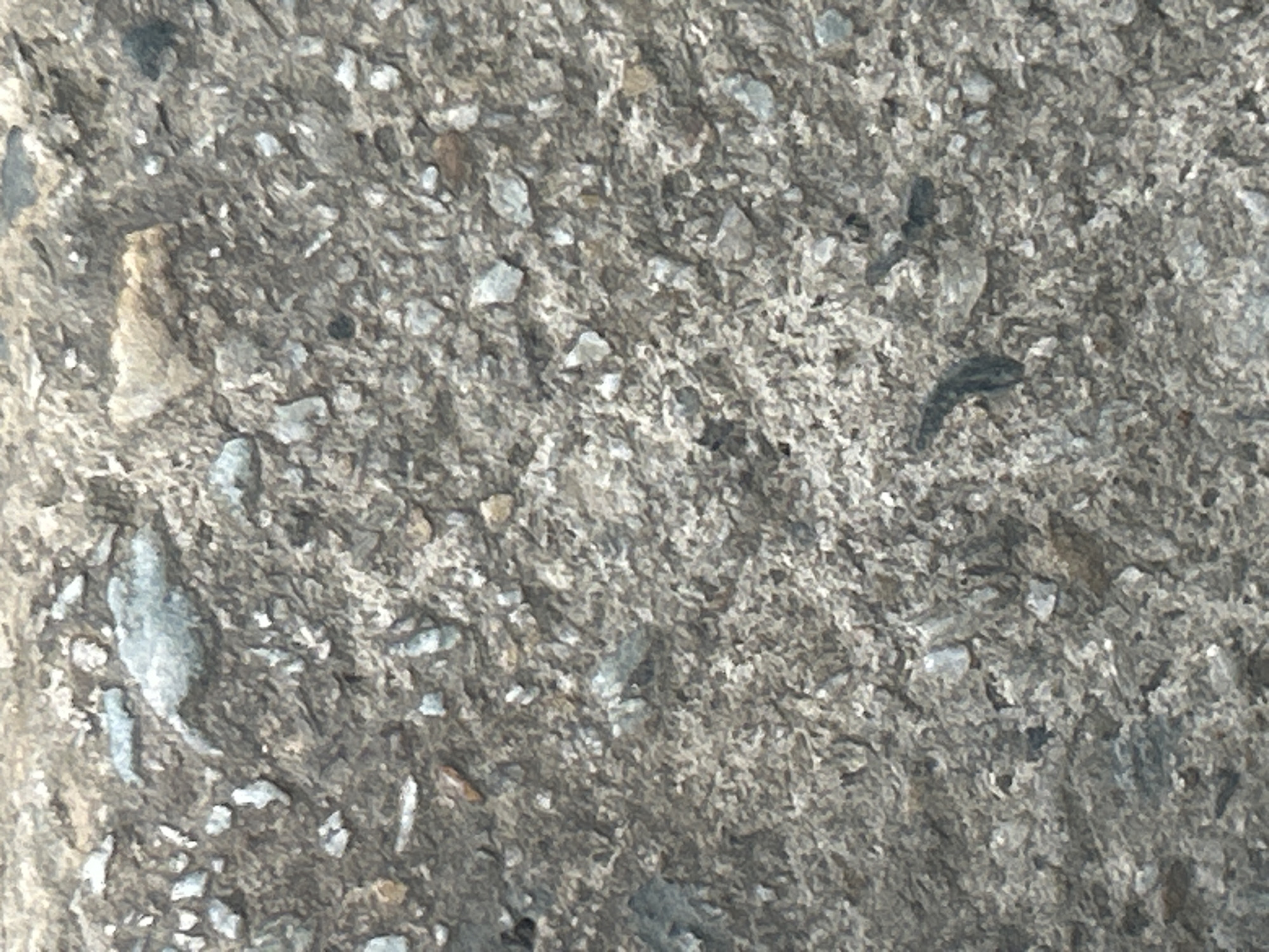 The image shows a close-up, textured view of a weathered concrete surface, possibly a wall or pavement. The concrete is a mix of gray cement and embedded small stones and pebbles of various shades of gray, white, and brown. Some larger fragments of rock are also visible, along with irregular pits and cracks in the surface, indicative of age and exposure to the elements. The overall impression is of a rough, granular, and somewhat eroded material. There are no people, animals, or discernible objects other than the concrete and its constituent materials. The location in Santa Ana, Venezuela, cannot be determined from the image itself, as it only depicts a generic material texture.