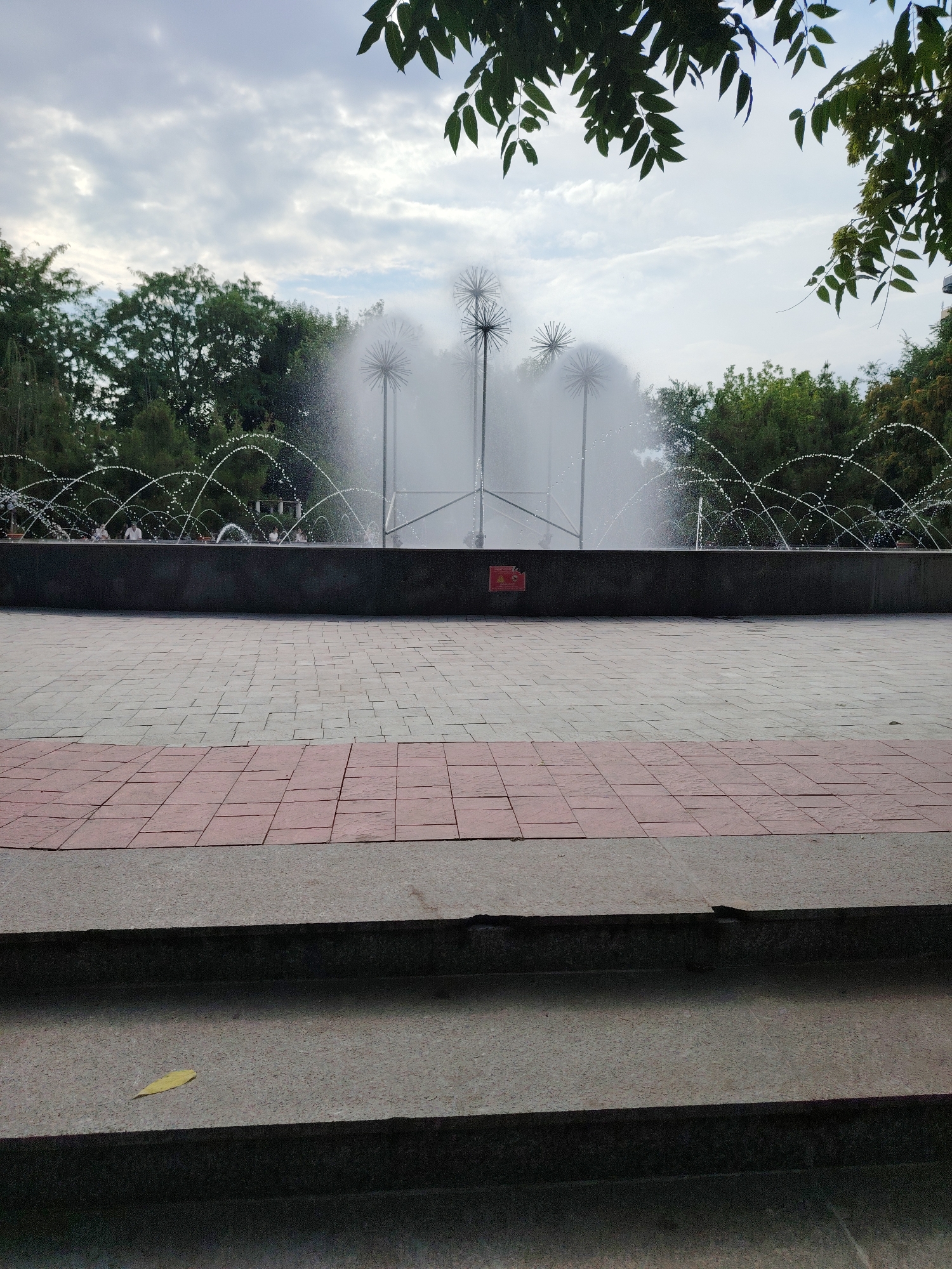 The image shows a wide shot of an outdoor fountain with arched water jets. The fountain is situated in a paved area with a dark, rectangular basin. Behind the fountain, lush green trees form a dense backdrop under a cloudy sky. The foreground features a walkway made of light gray and reddish-brown rectangular tiles, with a set of dark gray concrete steps leading down from it. A single yellow leaf rests on the second step from the bottom. Several spiky, metallic sculptures rise from the water of the fountain, creating a unique visual effect. In the distance, behind the fountain, several people are visible, appearing as small figures. The lighting suggests it is daytime, possibly late afternoon or early evening, with diffused light from the overcast sky. The overall scene conveys a sense of a public park or plaza, with a serene and somewhat tranquil atmosphere. A small red rectangular sign with white text is affixed to the side of the fountain basin, but the text is illegible.