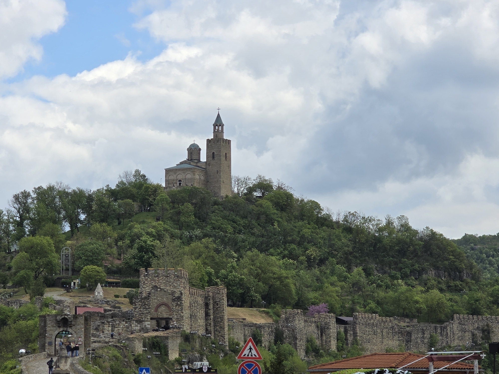 The image depicts a historical site located in Unknown, Unknown. A church with a tall tower and a dome stands atop a tree-covered hill. The church is constructed of light-colored stone. Below the church, there are remains of an old stone wall with arches and other architectural features. There are some people present on a walkway and stairs near the bottom left of the image. The sky is partly cloudy. There are some road signs visible near the bottom. The overall environment is lush and green.