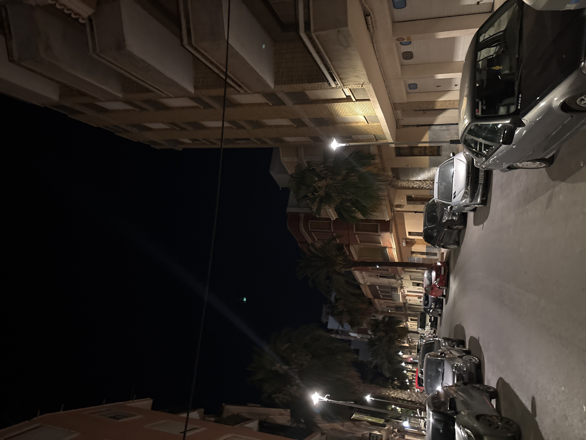 The image depicts a street scene at night in Picassent, Spain. The street is lined with multi-story buildings and parked cars. On the left side of the street, there is a peach-colored building with white-trimmed windows. Further down, palm trees are visible. On the right side, a tall, tan brick building with balconies is prominent. Several cars are parked along the street, including a silver sedan in the foreground on the right, and other vehicles of various colors and models filling the street. Streetlights illuminate parts of the scene. The sky is dark, indicating nighttime. There are no people visible in the image. The environment suggests a residential or mixed-use urban area.