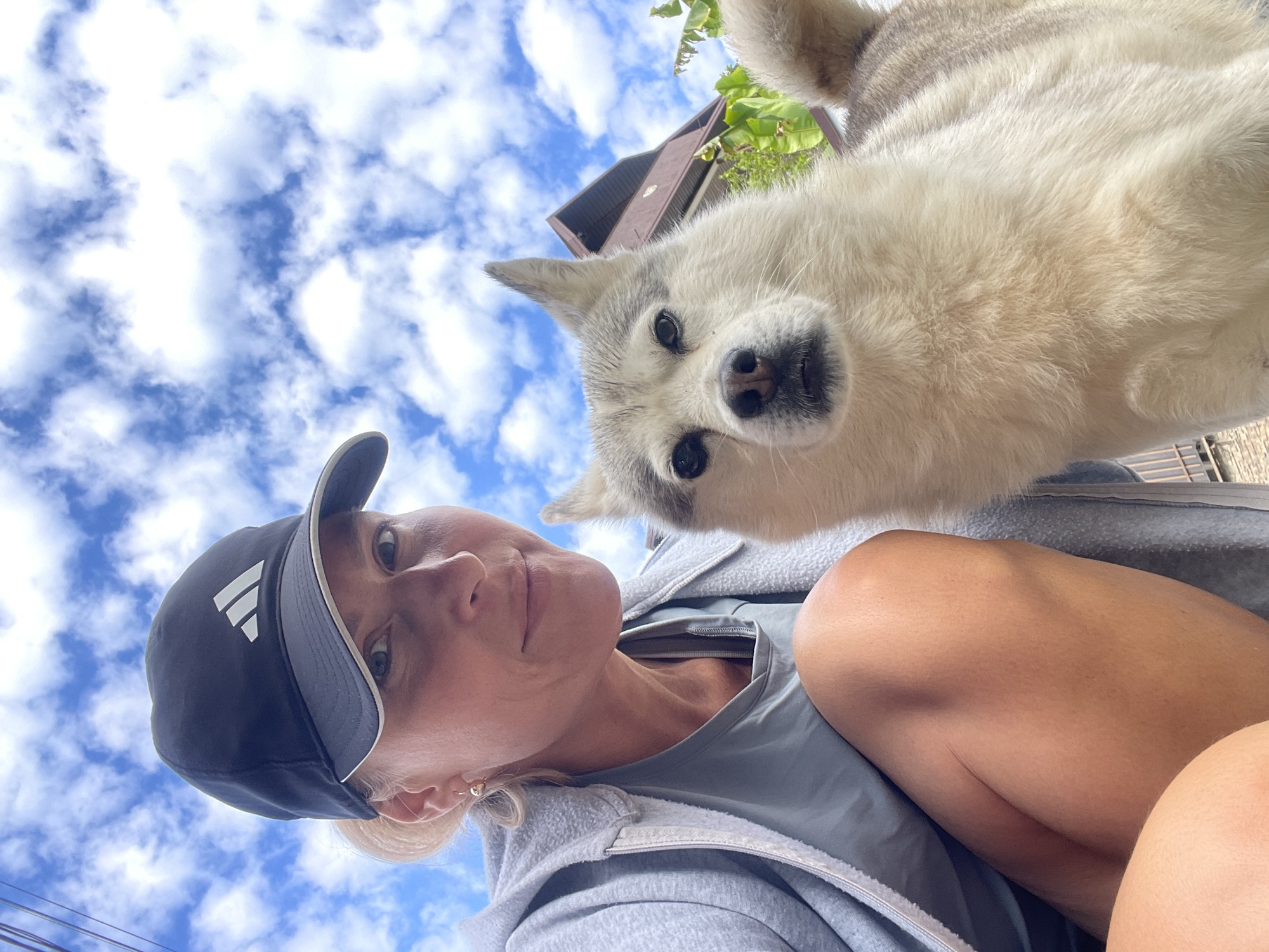The image shows a selfie taken from a low angle. A woman wearing a dark baseball cap is positioned in the lower left of the frame. Part of her upper torso and a portion of her leg are also visible. Above her and slightly to the right is a dog, possibly a husky or malamute, with its head angled down toward the camera. The background consists of a bright blue sky with scattered white clouds. A building is partially visible in the upper right corner of the frame. There is no information to confirm the location is Baranovka, Russia.