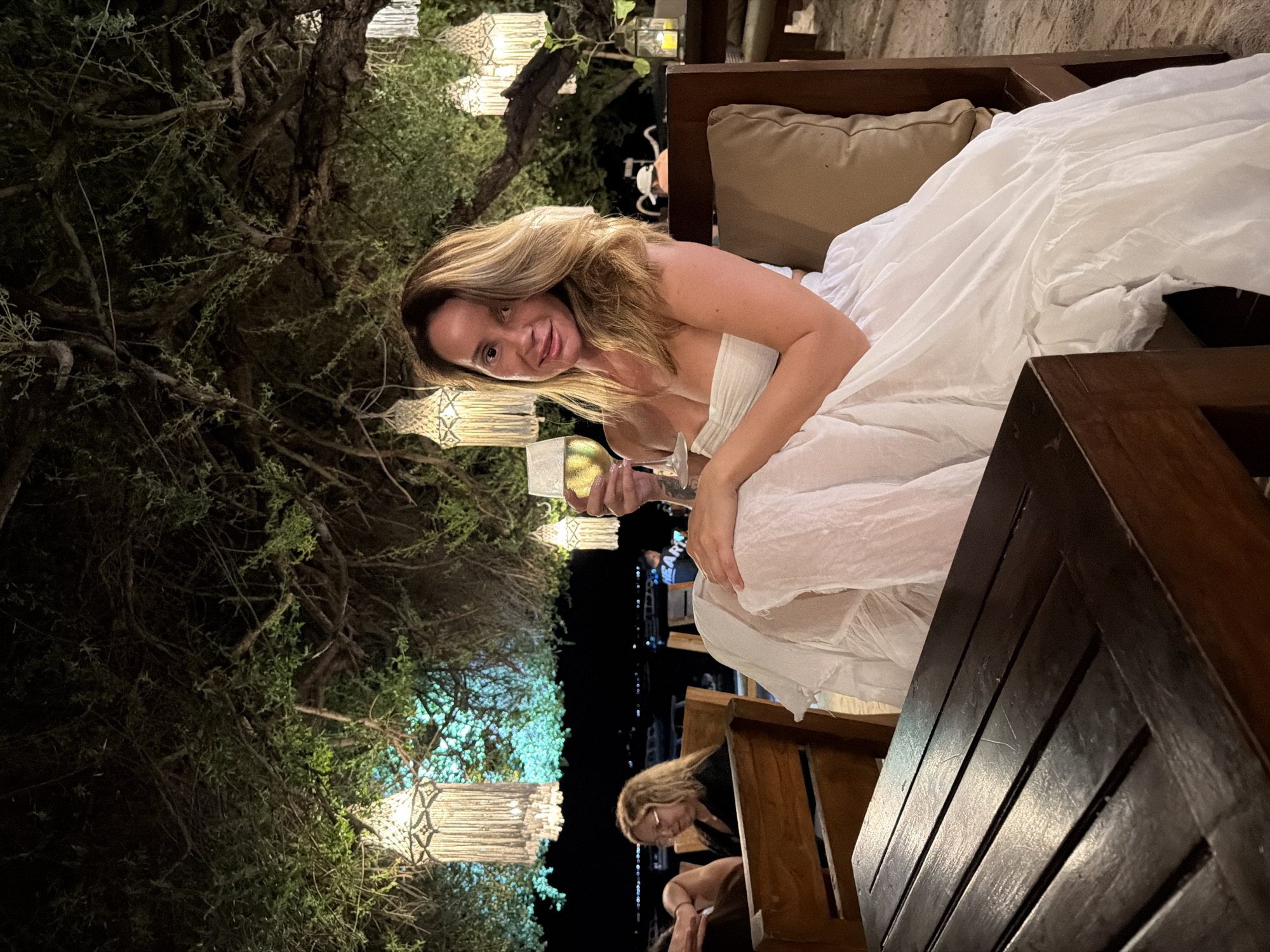 A woman with blonde hair is sitting at a table outdoors at night, holding a glass of white wine. She is wearing a white strapless dress. She is looking at the camera. There are trees with lights hanging from them in the background. There is a dark wooden table in the foreground. There are other people sitting at tables in the background. The location is a beach restaurant.