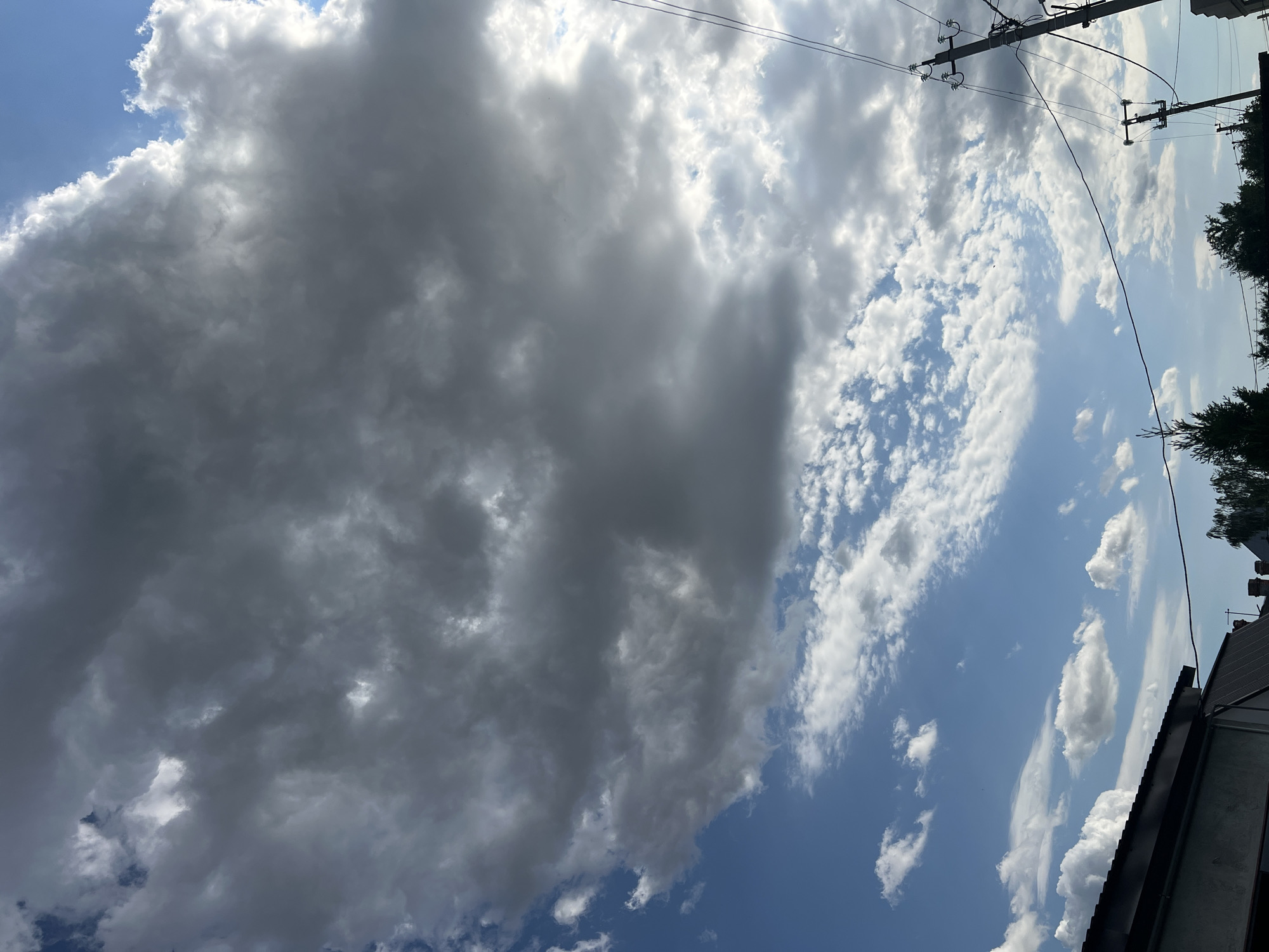 The image depicts a cloudy sky with a portion of rooftops and trees visible at the bottom. The sky is a mix of bright blue and dark gray clouds, with sunlight filtering through in some areas. The clouds appear to be dense and textured, suggesting a dynamic weather system.
In the lower part of the frame, the dark roof of a building is visible on the left, with some trees and what appear to be chimneys or antennae on other structures further to the right. A utility pole with wires is also prominent on the right side of the image. The scene is shot from a low angle, looking upwards towards the sky.
This image was taken in Tiraspol City Council, Moldova.