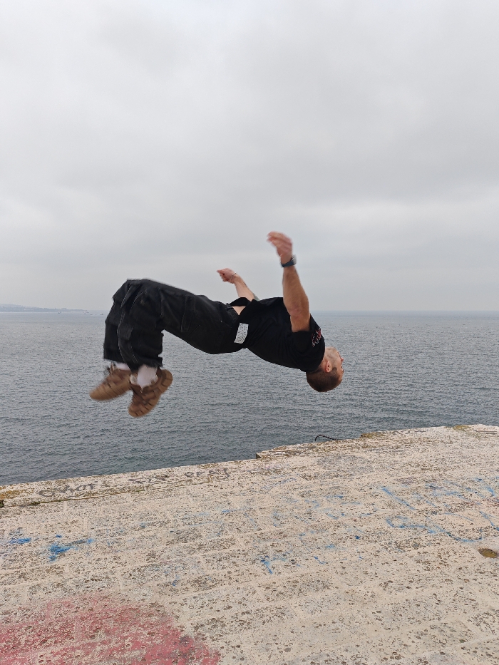 A male individual is suspended horizontally in mid-air, performing an acrobatic backflip over a concrete sea wall in Varna, Bulgaria. He is inverted, with his head pointing towards the ground and his body arced, arms bent and raised for momentum. He is dressed in a black short-sleeved shirt, dark-colored cargo pants, and light brown footwear, possibly sandals, worn over white socks. A dark wristwatch is visible on his left wrist. The concrete surface below him is weathered and marked with faded red and blue graffiti. Beyond the concrete, the dark blue-grey waters of the Black Sea are calm with subtle ripples. The horizon is visible, and a faint landmass or coastline stretches into the distance on the left. The sky above is uniformly overcast and grey.