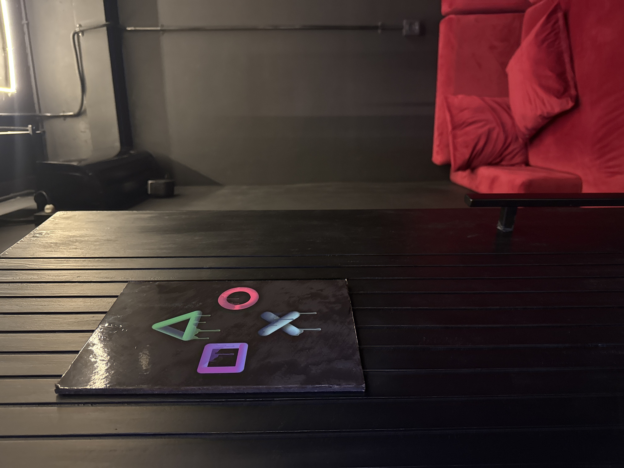 The photograph shows a black slatted table with a square tile featuring a PlayStation logo. The tile sits on the left side of the table, showing the iconic PlayStation symbols: triangle, circle, cross, and square, each in a different color. In the background is a red, velvet sectional couch with large pillows. The couch is situated against a dark gray wall. To the left of the couch, there's a dark, ambiguous object, possibly a piece of equipment, near a wall with pipes. The scene appears to be indoors, possibly in a lounge or game room. There is no explicit information about the location being in Kuta Selatan, Indonesia.