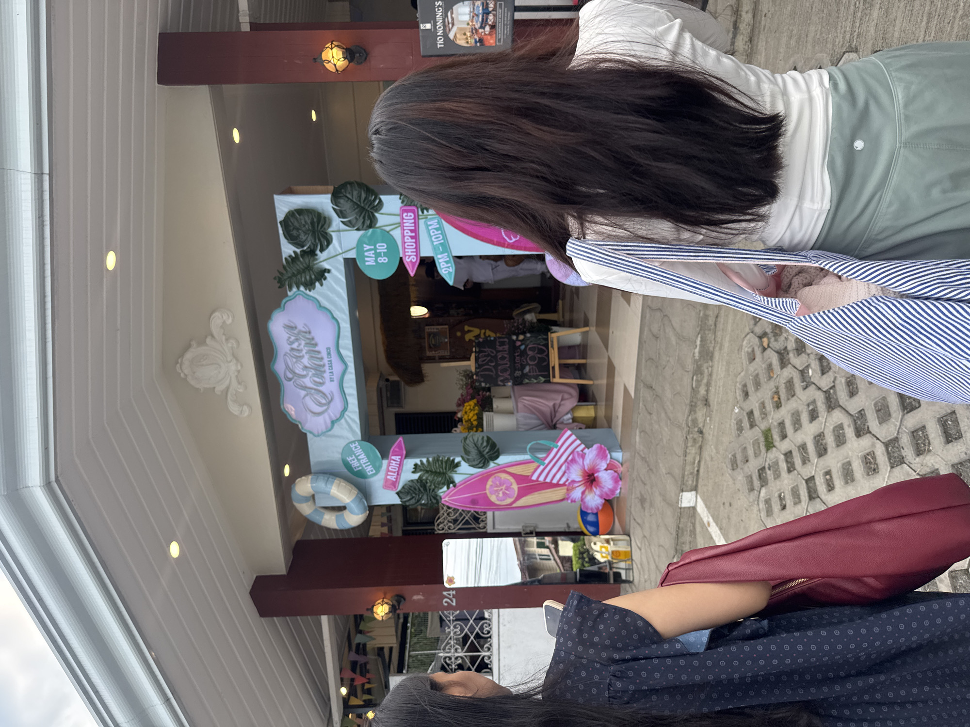 Two women are visible, standing in front of "Casa Cinco," a shopping event location in Davao City, Philippines. One woman, facing forward, has long, dark hair and is wearing a white top and light green bottoms. She holds a striped bag. The other woman, closer to the camera, is wearing a dark blue patterned top and carries a burgundy-colored purse. A sign reads "Casa Cinco" and also advertises the shopping event, "May 8-10, 2PM-10PM." Tropical decor is visible, including surfboards, flowers, and inflatable rings. A smaller sign reading "Tío Noning's" is visible above the storefront. The address "24" can also be seen.