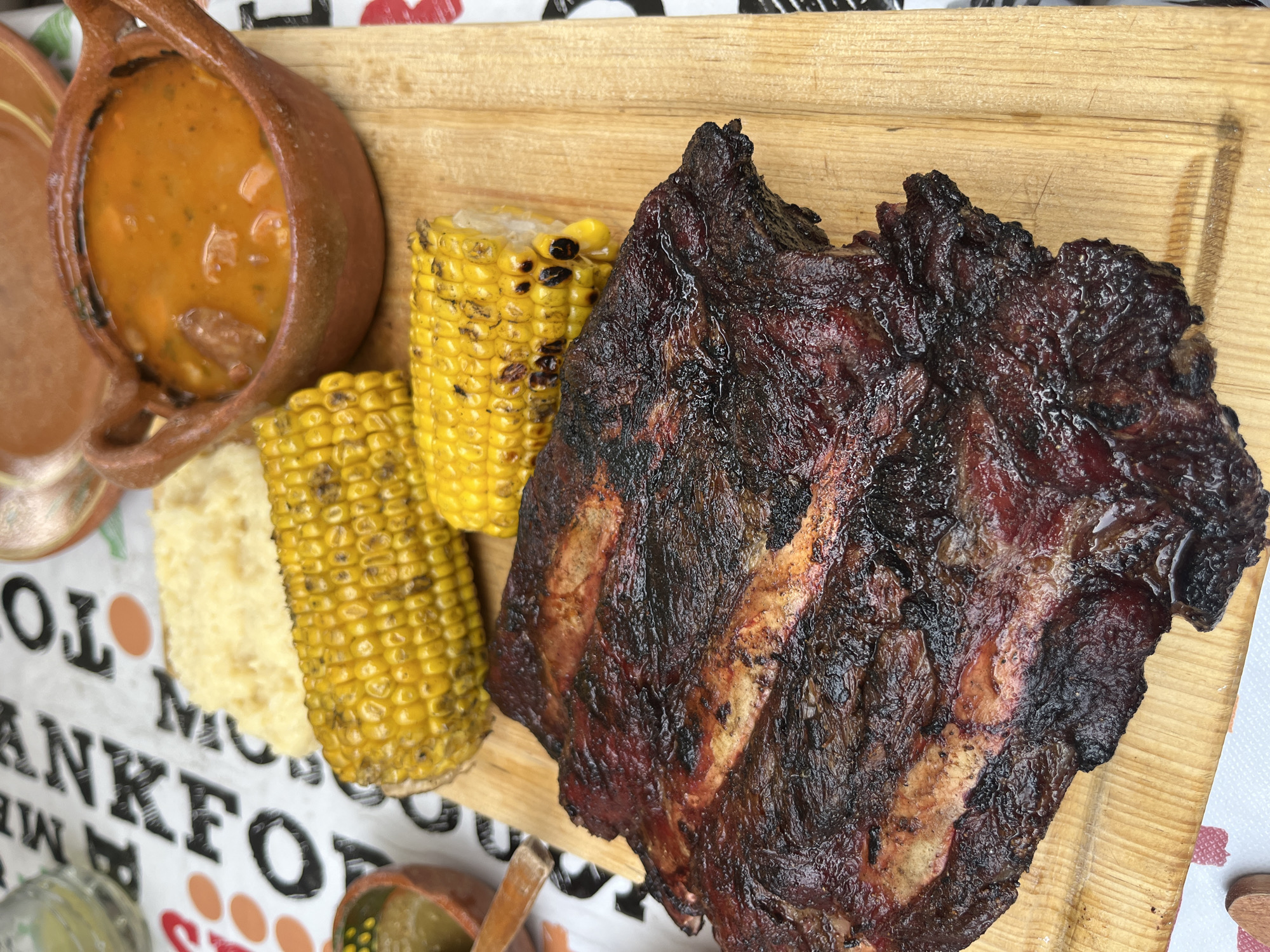 The image shows a wooden platter with a hearty meal. Featured are a rack of ribs with a dark, caramelized crust, two ears of grilled corn, and a small terracotta pot filled with a stew. Beside the corn is a small portion of white mashed or puréed food. The platter rests on a surface covered with a patterned tablecloth. The scene evokes a sense of rustic dining, possibly outdoors. The location given is San Francisco Totimehuacán, Mexico.