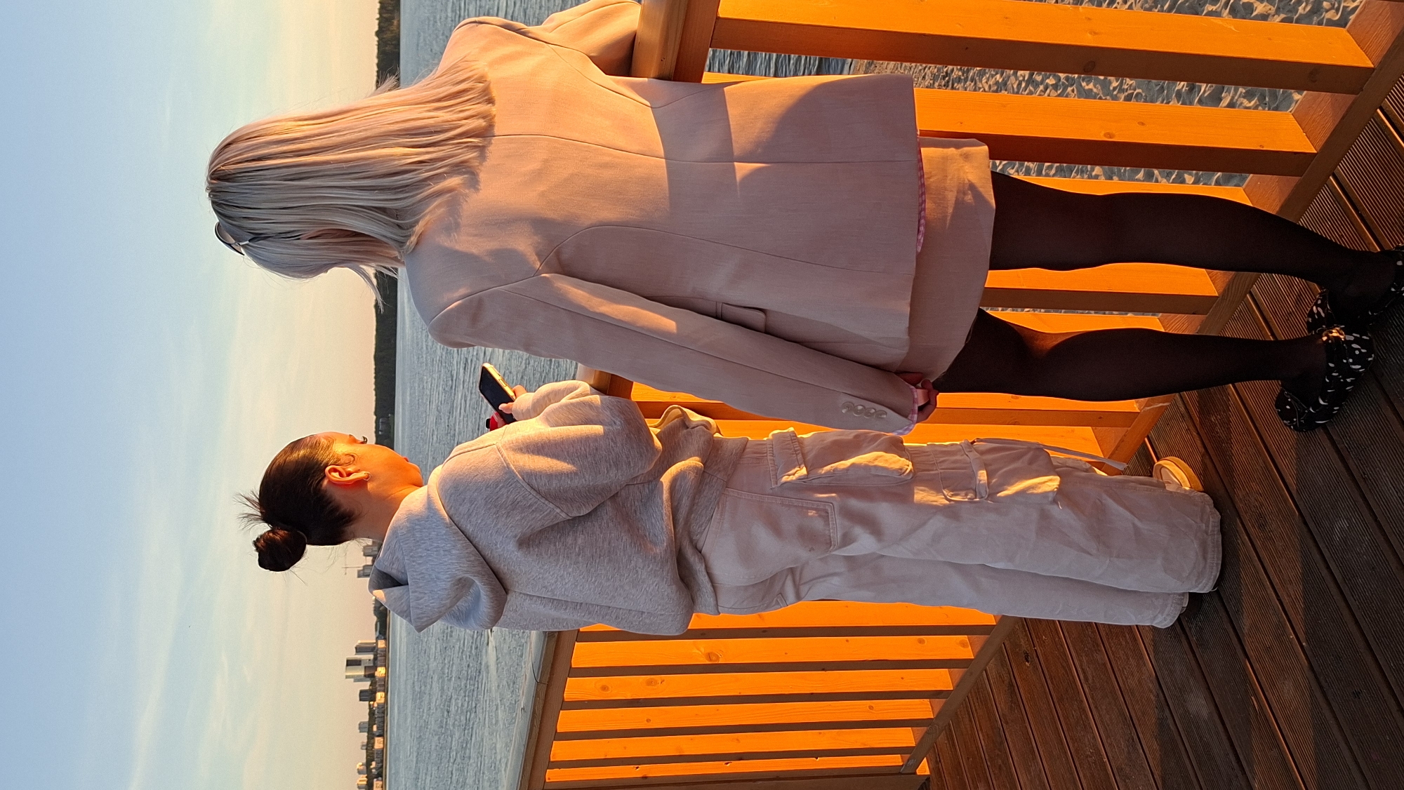 Two individuals stand on a wooden boardwalk near a body of water, with the city skyline of Tallinn, Estonia, visible in the distance. The sun is low in the sky, casting a warm, orange glow on the scene. The person on the left, facing away from the camera, has their hair in a bun and is wearing a light-colored hoodie and cargo pants. They are holding a smartphone. The person on the right, also facing away, has blonde hair and is wearing a light-colored blazer, a skirt, and black tights. They are standing near a wooden railing. The boardwalk appears to be made of dark wood planks, and there is sand visible beyond the railing to the right.