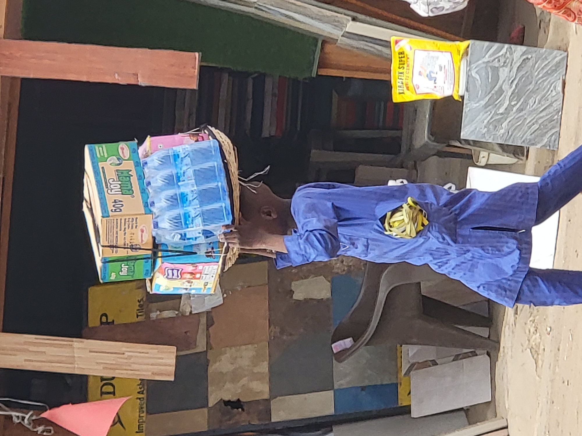 A man in a blue robe carries a woven basket of bottled water and other items on his head in an outdoor setting, likely a marketplace. The man's robe is secured around his waist with a yellowish-green tie. Various products like tiles, detergent (Mama Joy), and cement (Kiala Fix Super Anti-Cement) are visible in the background. The location appears to be a commercial area, possibly in Maiburiji, Nigeria, given the context and the items for sale. The floor is tiled in a checkered pattern of various colors. A chair is situated behind the man, adding to the impression of an active marketplace or trading environment.