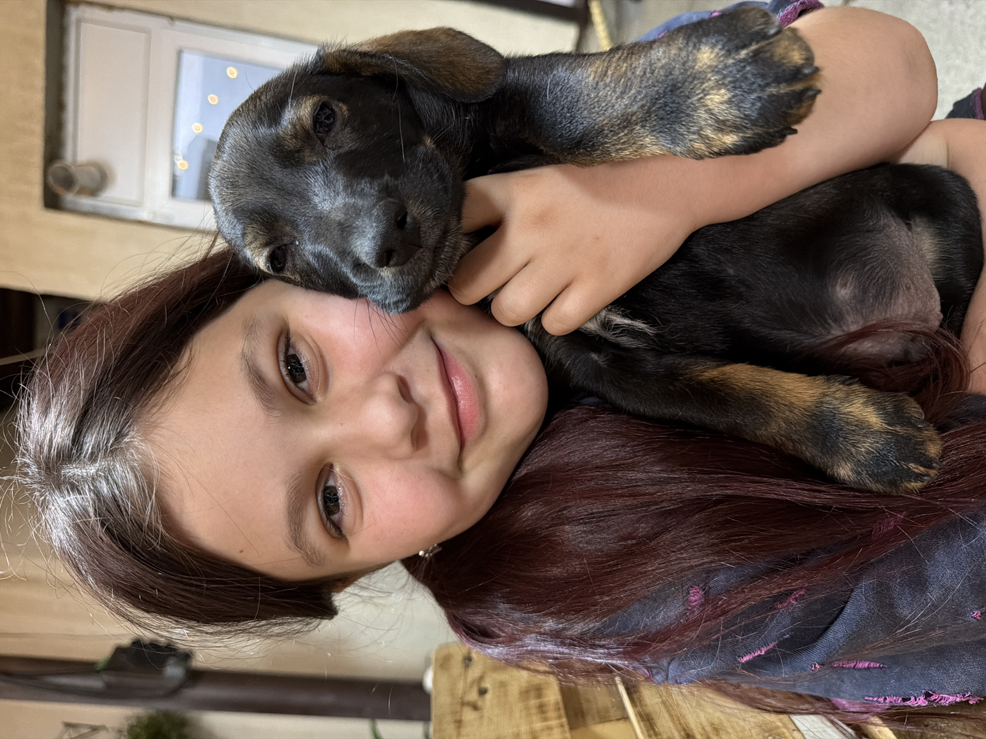 The image depicts a young girl holding a black and tan puppy. The girl, with long, dark, reddish-brown hair, smiles at the camera, her head tilted. She is wearing a dark-colored top with some pink accents. The puppy is nestled against her chest, its head resting on her cheek. The puppy has dark fur with tan markings on its legs and face. They appear to be indoors, with a wooden structure visible to the left and a blurred background that suggests a building with a window. No specific landmarks indicating the city of Tuapse, Russia, are visible in the image.