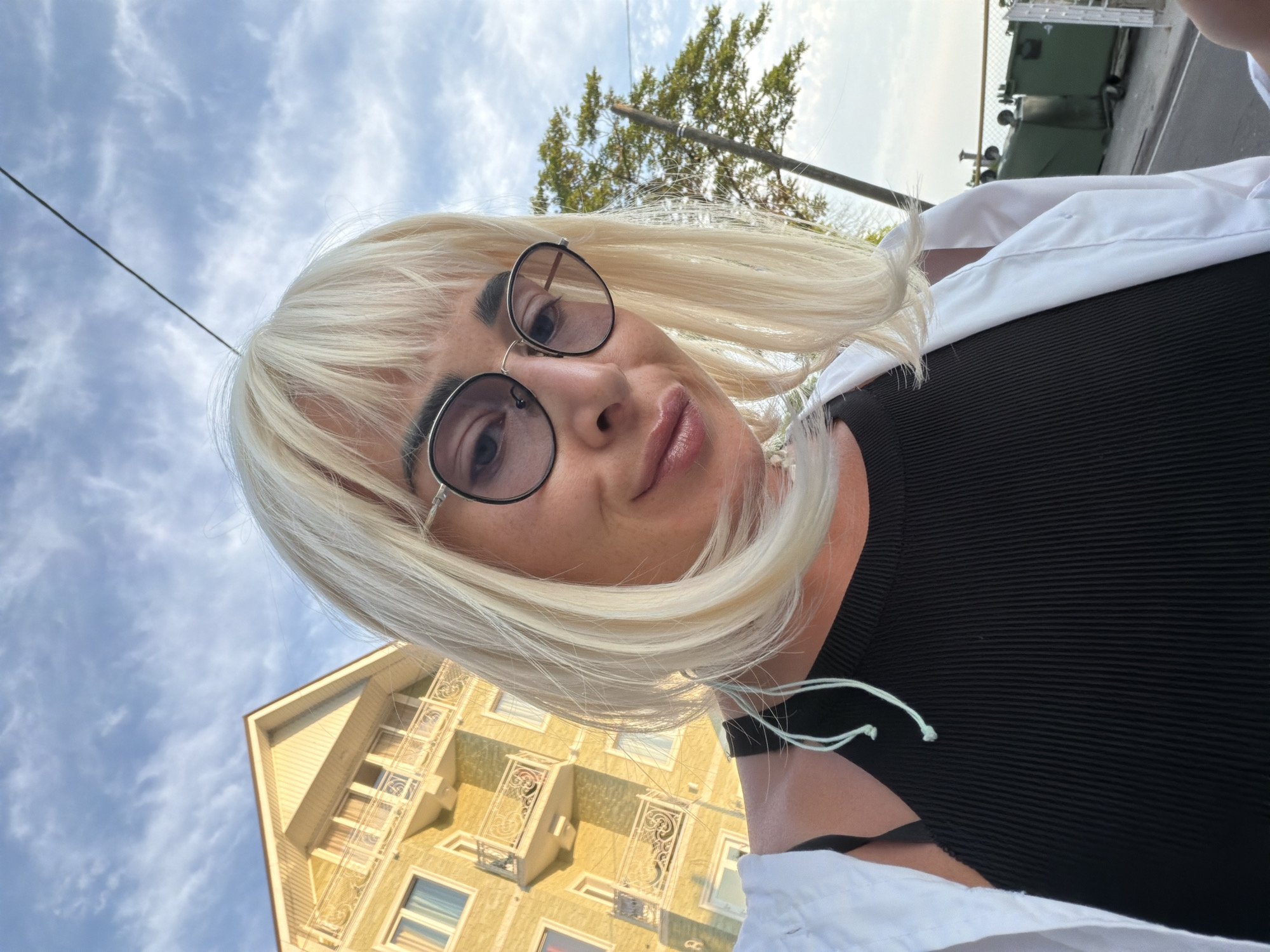 This is a low-angle, close-up, selfie-style photograph taken outdoors. The subject is a person with blonde hair, wearing round-framed glasses and a black ribbed top, with a white jacket or shirt draped over their shoulders. They are looking towards the camera with a slight smile.
In the background, a yellow building with decorative balconies and windows is visible against a partly cloudy sky. A tree with green foliage is also in the background, along with a utility pole and a power line. Further in the distance, two green dumpsters and part of a fence can be seen. The lighting suggests it is daytime, likely late afternoon due to the soft light and shadows. The overall mood is casual and relaxed. There is no visible text in the image. The context provided indicates the location is Sochi, Russia.