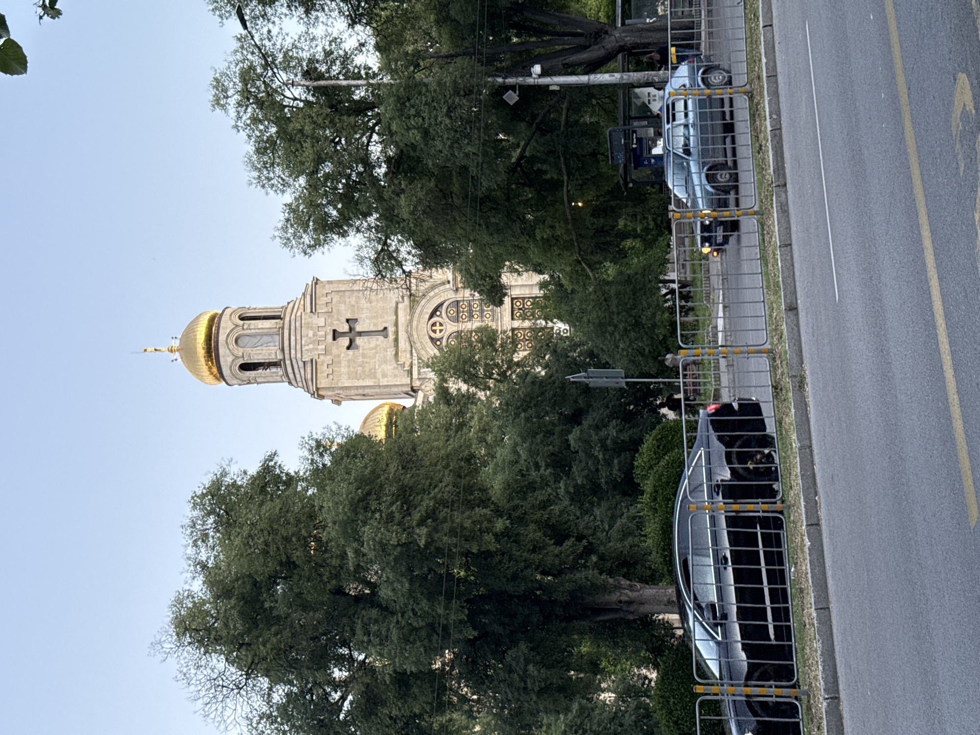 The image shows a wide shot of a street scene in the city of Unknown, Unknown. In the background, a large church with golden domes and a cross is visible, surrounded by lush green trees. In the foreground, a black car is parked behind a metal fence with yellow and silver poles. To its right, a grey station wagon is also behind the fence, with its headlights on. The street in front of the fence is paved with asphalt and has yellow lane markings. A bus stop with a bus shelter is visible behind the grey car. The sky is clear and blue.