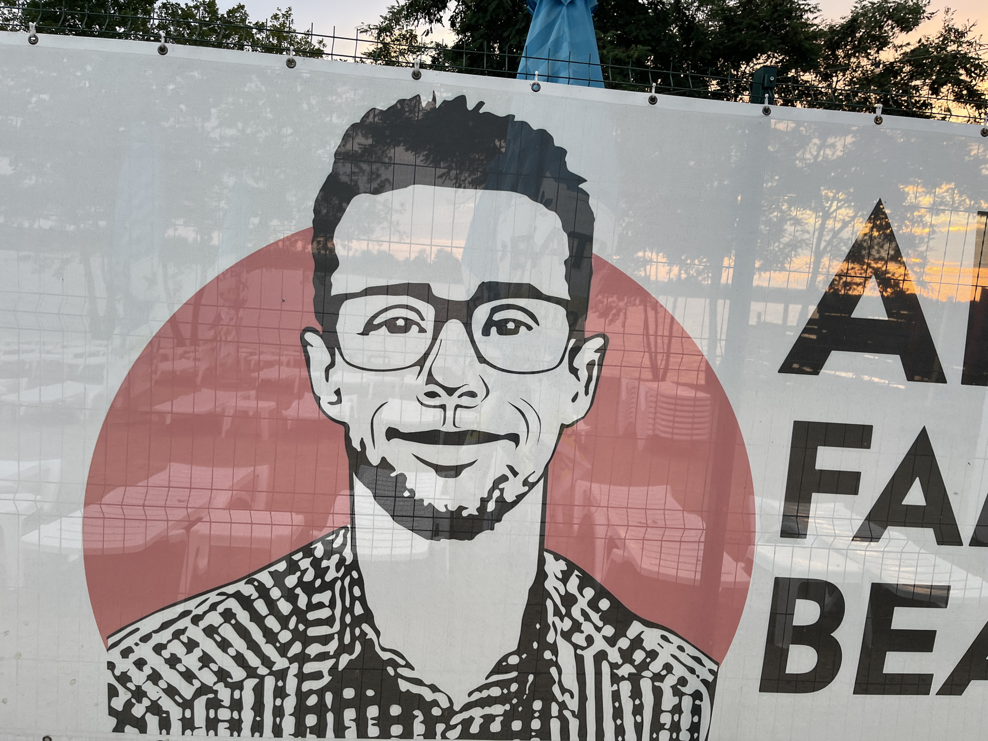 A large banner displays a stylized black and white portrait of a man wearing glasses and a patterned shirt. A large red circle frames the left side of his face. To the right of the portrait, large black letters form the beginning of words: "AI", "FA", "BEA". The banner appears to be made of a mesh material, allowing the background scene to be visible through it. This background includes trees with dappled sunlight filtering through, suggesting it is daytime. The sky is visible in the upper portion of the image, with a warm, sunset-like glow. In the background, beyond the banner, a chain-link fence and trees are visible, and a blue tarp or flag can be seen near the top. The banner is attached to a fence with metal grommets. The overall impression is that of an outdoor event or display, possibly a political or promotional banner given the partial text. The lighting indicates it is either early morning or late afternoon.