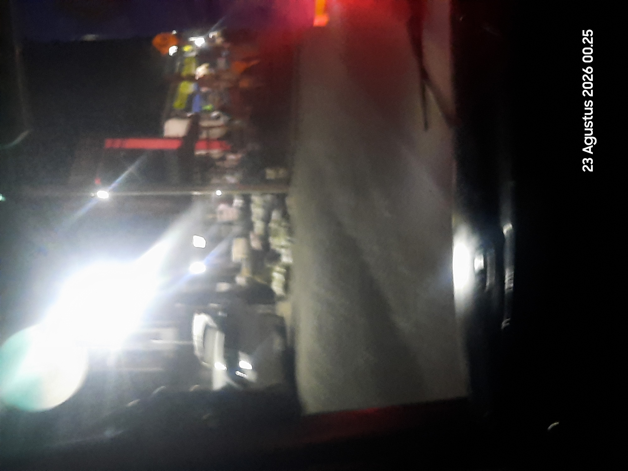The image depicts a nighttime scene, possibly taken from inside a vehicle, looking out onto a street in Sukokerto, Indonesia. A bright light source, likely a streetlamp or a large bulb, dominates the upper left portion of the frame, casting strong glare. In the middle ground, a light-colored car is visible, parked or moving slowly on the road. To the right of the car, a market or street vendor area is suggested by blurred figures and various stalls or displays illuminated by artificial lights. The ground appears to be asphalt or pavement. The bottom of the image shows the interior of a vehicle, possibly a dashboard or the area around the windshield. The timestamp "23 Agustus 2026 00.25" is present in the lower right corner, indicating the date and time. The overall ambiance is dark, with high contrast due to the lighting conditions.