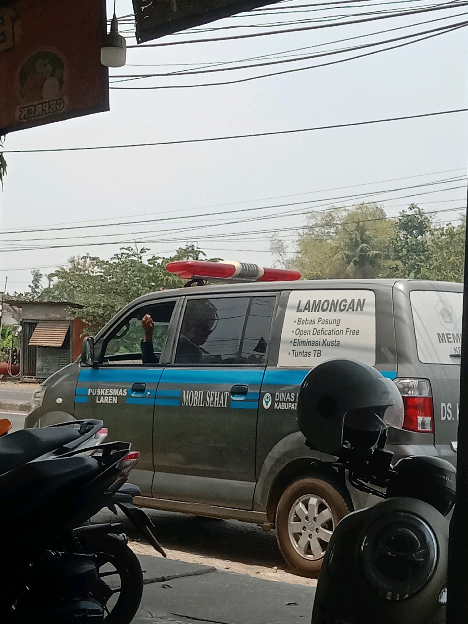 The image depicts a street scene in Plosowahyu, Indonesia, with a mobile health vehicle prominently featured. The vehicle, a dark-colored van with blue stripes, bears text identifying it as belonging to "PUSKESMAS LAREN" and "MOBIL SEHAT." A decal on the side of the vehicle lists objectives: "LAMONGAN," "Bebas Pasung," "Open Defication Free," "Eliminasi Kusta," and "Tuntas TB." A person's arm is visible through the open driver's side window. In the foreground, two dark-colored motorcycles are parked. The background shows a building with a red-topped light fixture and overhead power lines against a hazy sky.