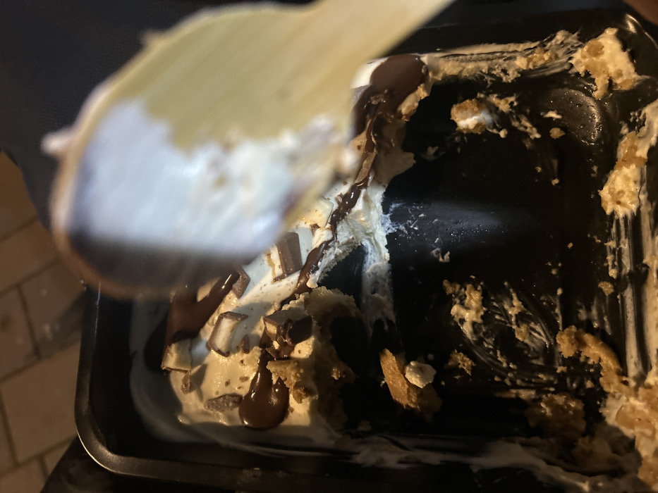 The image displays a close-up view of a partially consumed dessert contained within a black, rectangular plastic vessel, located in Ostwald, France. The dessert is composed of a light-brown crumbled biscuit base, a substantial layer of white or off-white cream, and is topped with dark brown chocolate sauce drizzles and scattered, small, rectangular segments of chocolate bar with a light filling. Visible within the excavated portions of the dessert are dark, round cookie pieces. Approximately half to two-thirds of the dessert has been removed from the container. A light-colored, out-of-focus spoon, possibly wooden, is positioned in the upper right, laden with a scoop of the white cream, indicating active consumption. The scene is dimly lit, with a blurry background showing what appears to be a paved outdoor surface and dark fabric.