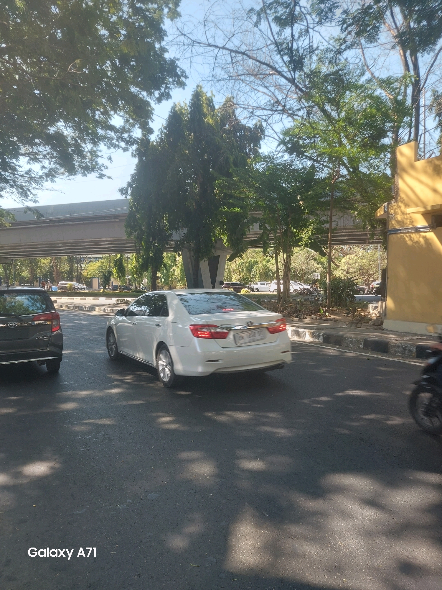 An exterior, eye-level, medium shot captures a street scene in Makassar, Indonesia, on a sunny day. In the foreground, a white sedan is traveling on the right lane of a road, with its rear facing the camera. To its left, a dark SUV is also in motion. In the background, an elevated road or overpass runs horizontally across the frame, supported by concrete pillars. Lush green trees line the sides of the street and frame the elevated structure. A yellow building with a dark awning is visible on the right side of the image. Shadows from the trees are cast on the asphalt road, indicating the bright sunlight. The overall impression is a typical urban environment with traffic, infrastructure, and vegetation.