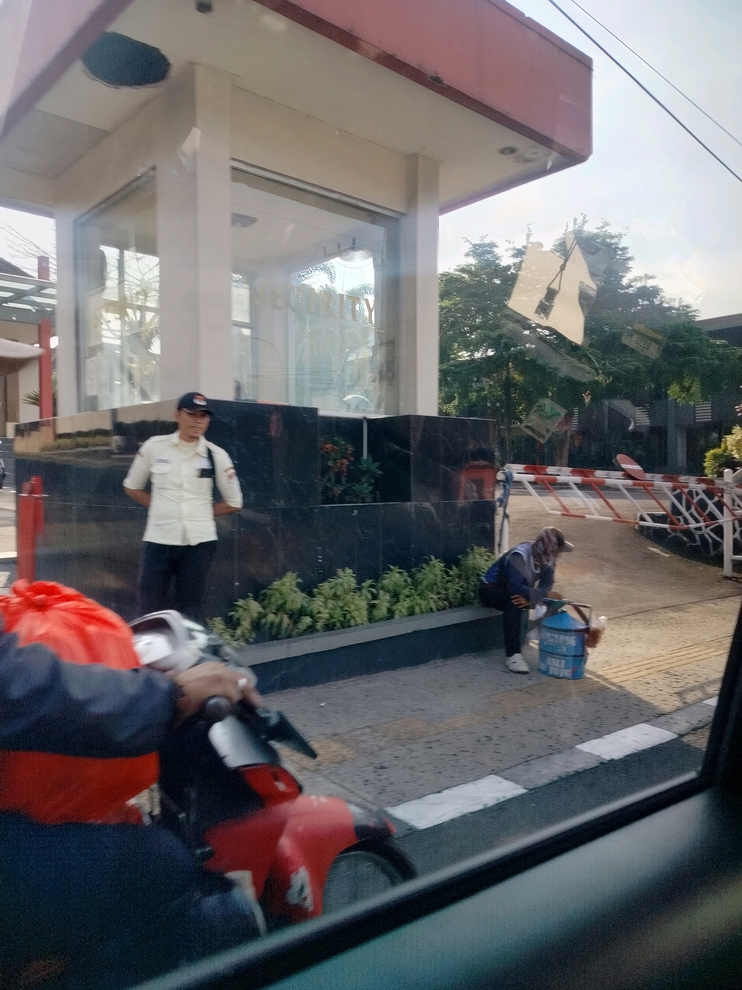 In Cisarua, Indonesia, a security guard in a white uniform and dark pants stands at attention in front of a glass-enclosed building with a red-trimmed roof. To the right, a person wearing a headscarf and a dark blue jacket sits on the ground next to a large blue bucket with white lettering. A red and white striped barrier gate is visible behind them. In the foreground, a person wearing a dark jacket over a bright red garment is seated on a red and silver motorcycle, with their hand on the handlebars. Green foliage lines the walkway in front of the building, and trees are visible in the background, under a bright sky.