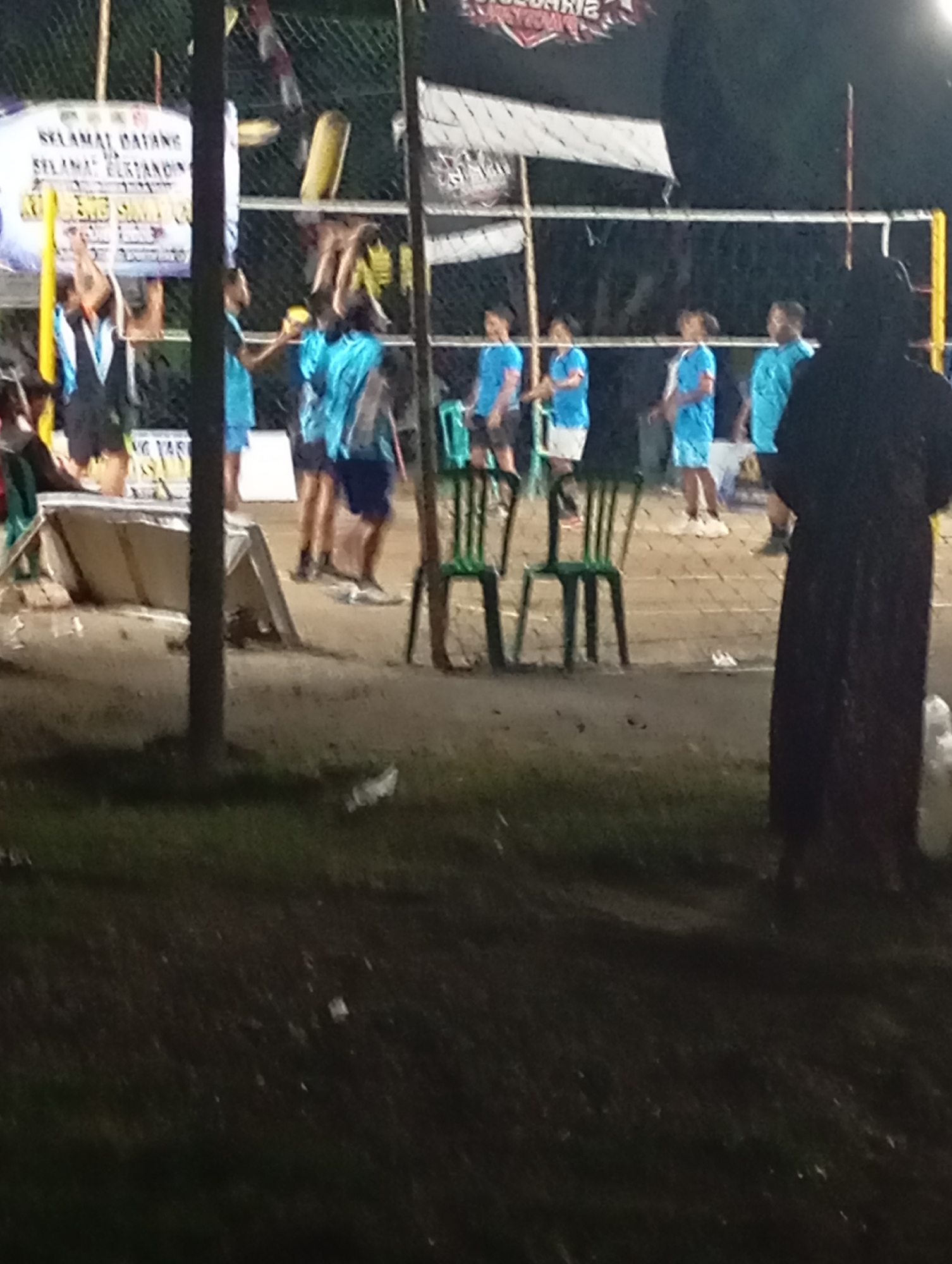 The image shows an outdoor volleyball court at night in Pacitan, Indonesia. A banner with text, including "SELAMAT DATANG" and "SELAMAT BERMAIN", is visible on the left side of the court. Several people, dressed in blue and black athletic wear, are present on and around the court, suggesting a game or practice is taking place. Some individuals are actively participating in the game, while others are observing. The court is lit, casting shadows and highlighting the sandy surface. Green plastic chairs are placed on the side of the court. The overall scene depicts a casual sporting event in an open-air venue.