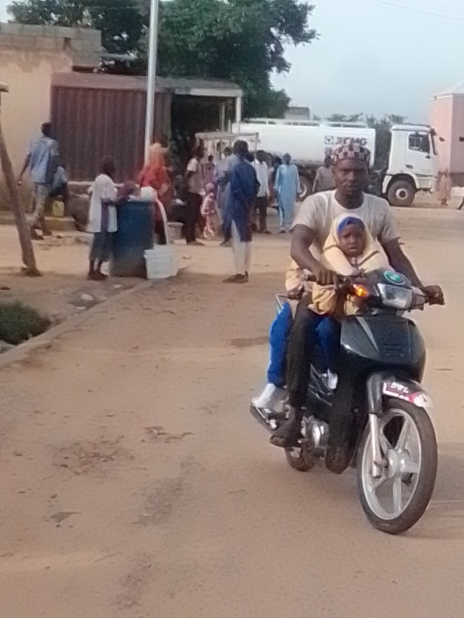 A man and a child are riding a motorcycle on a dirt road in Galadima, Nigeria. The man is driving the motorcycle, and the child is sitting in front of him. Both are wearing traditional African clothing. In the background, there are several buildings and a water truck. The sky is overcast.
